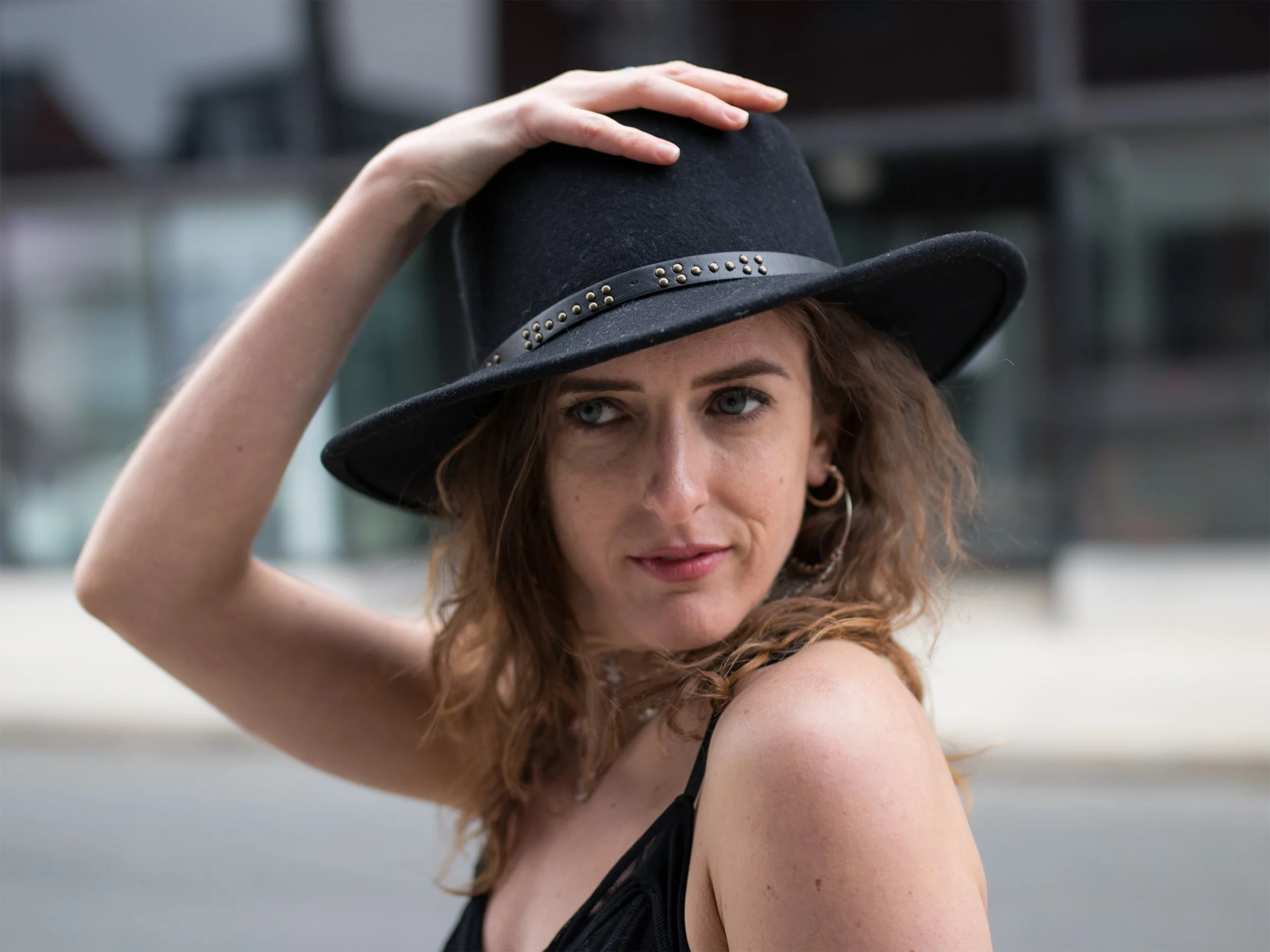 One hand rests on her black hat, a half-smile at her lips. Soft city light gives the portrait an ease that feels spontaneous.