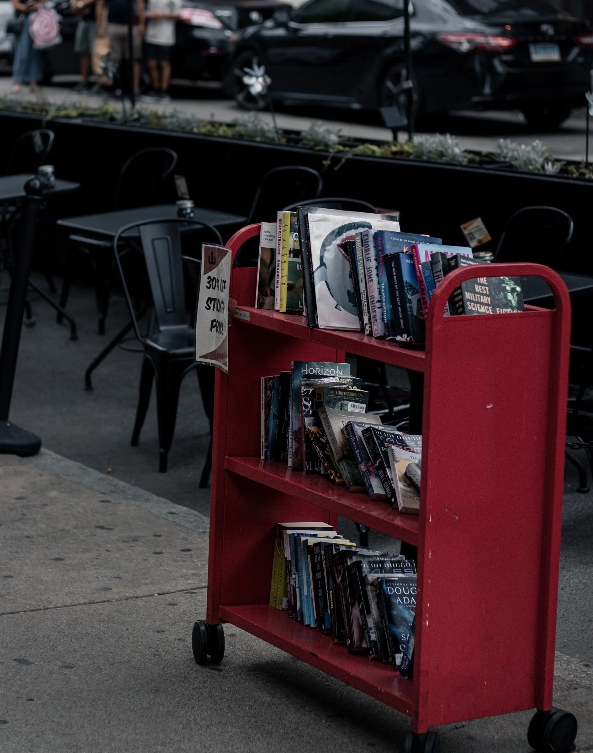 A worn red cart holds secondhand books beside an outdoor café.