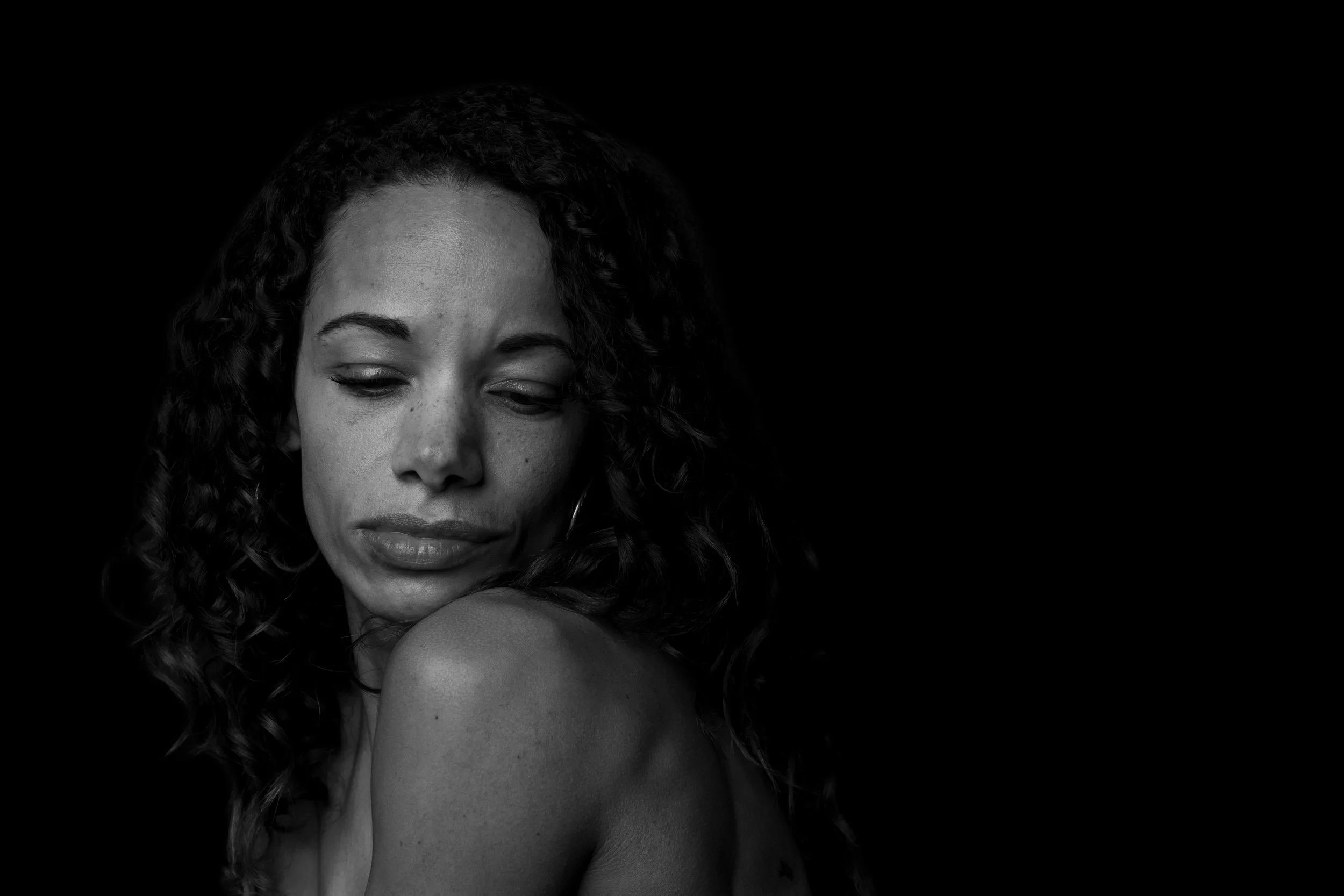 Her gaze falls over a bare shoulder, curls framing her face. The monochrome portrait draws attention to a moment turned inward.