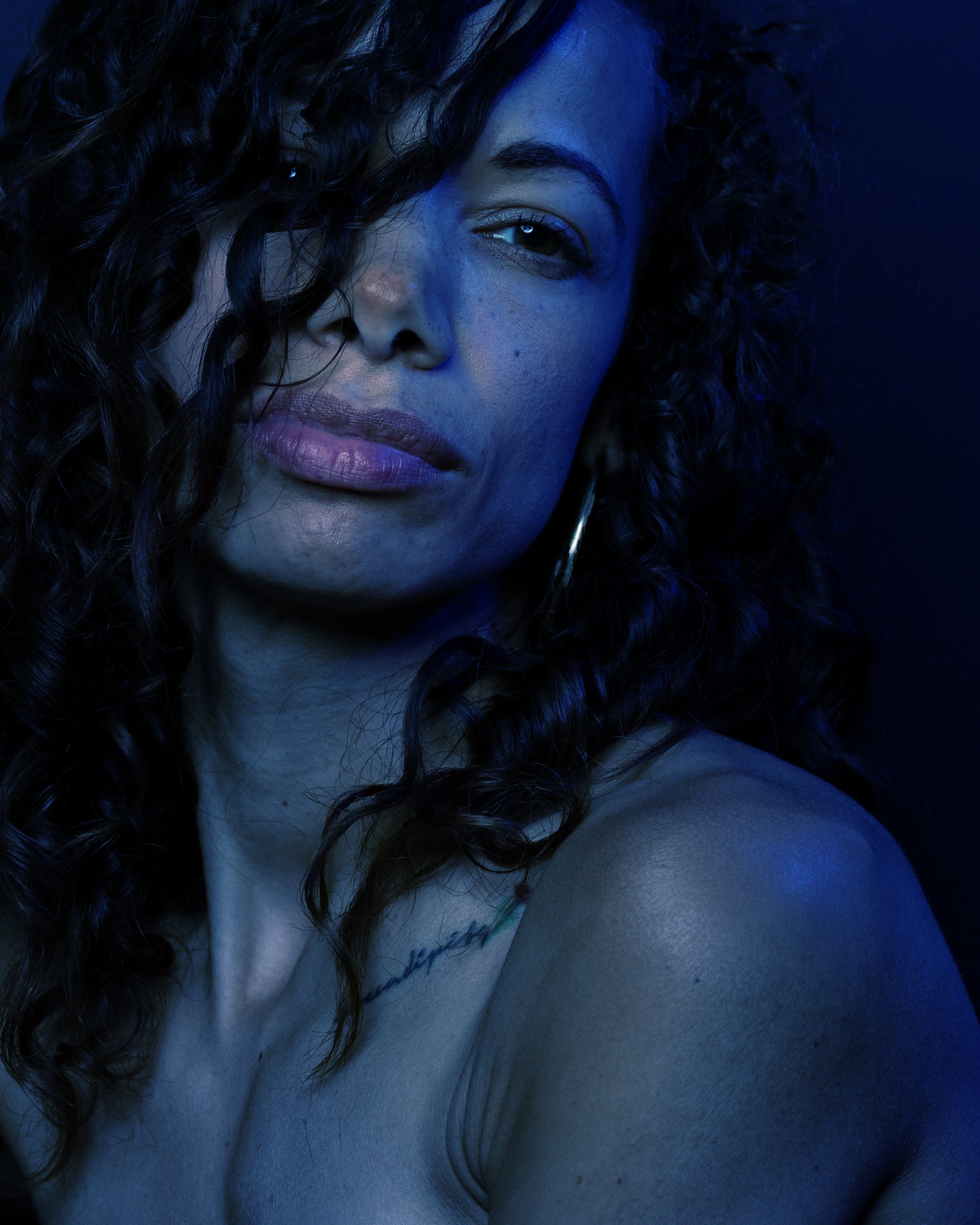 Blue light falls across her face and loose curls. Her unwavering expression brings tension to a portrait that feels almost within reach.