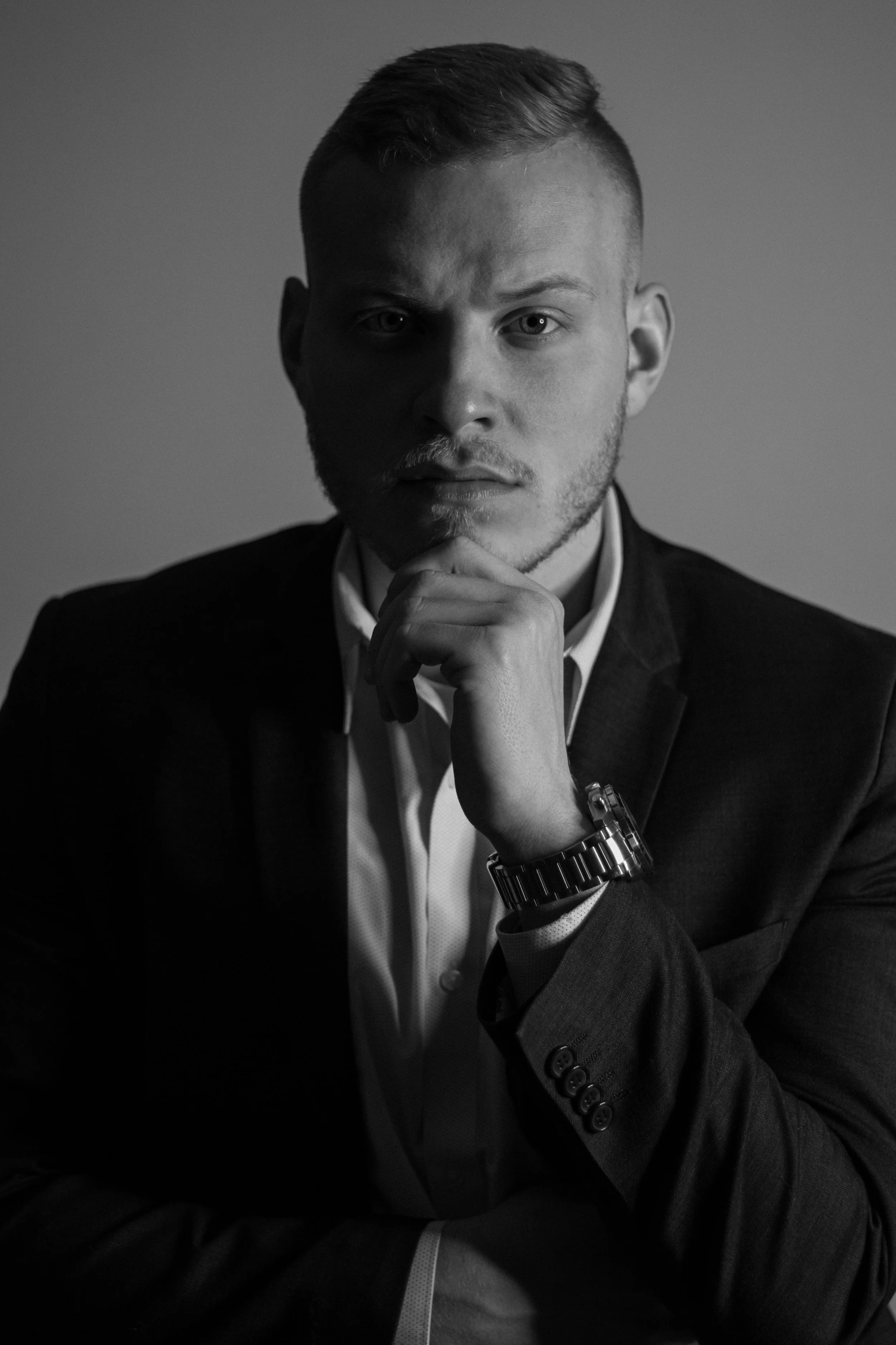 He rests his chin on one hand and looks directly at the camera, wearing a dark suit and a metal watch in a monochrome portrait.