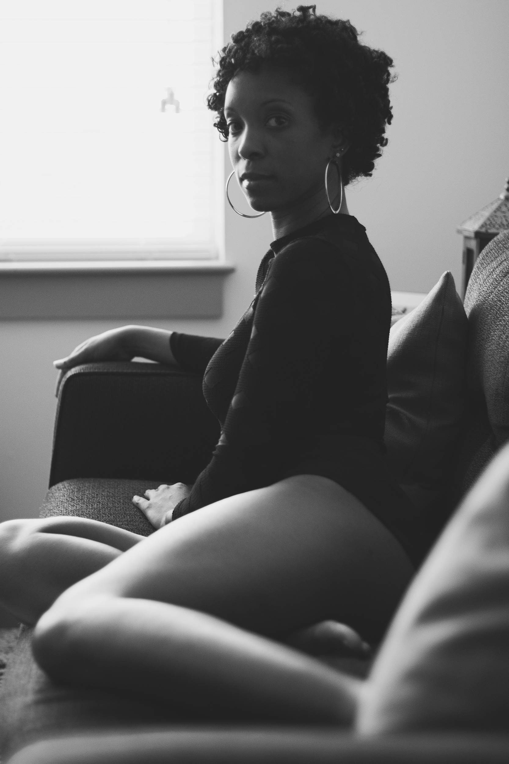 Seated beside a bright window, she turns back toward the camera. Soft light catches her curls and earrings while the monochrome shadows hold the intimacy of her gaze.