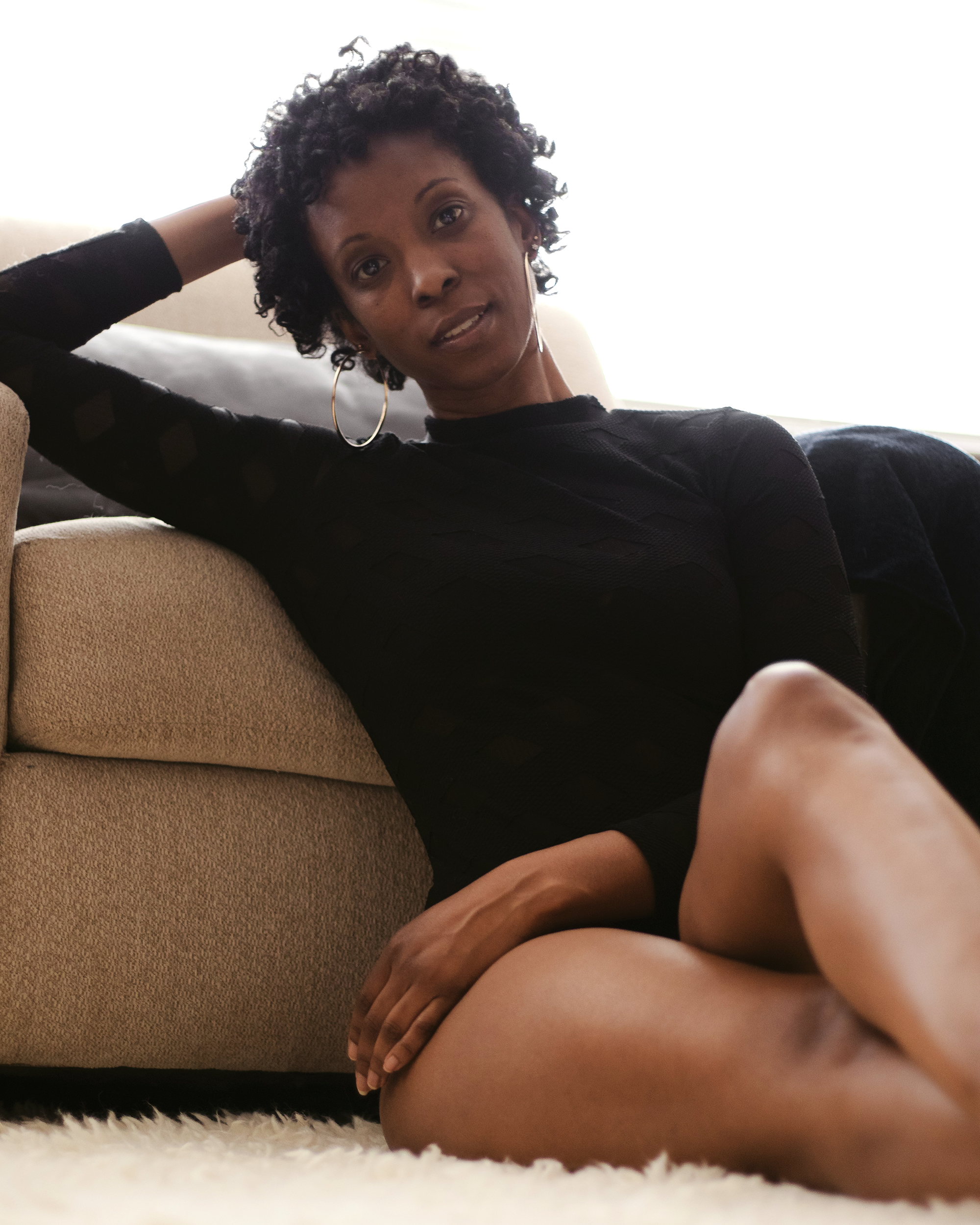 Seated low beside the chair, she looks directly toward the camera. A relaxed gesture, warm skin, and the soft foreground give the portrait a sense of presence and closeness.