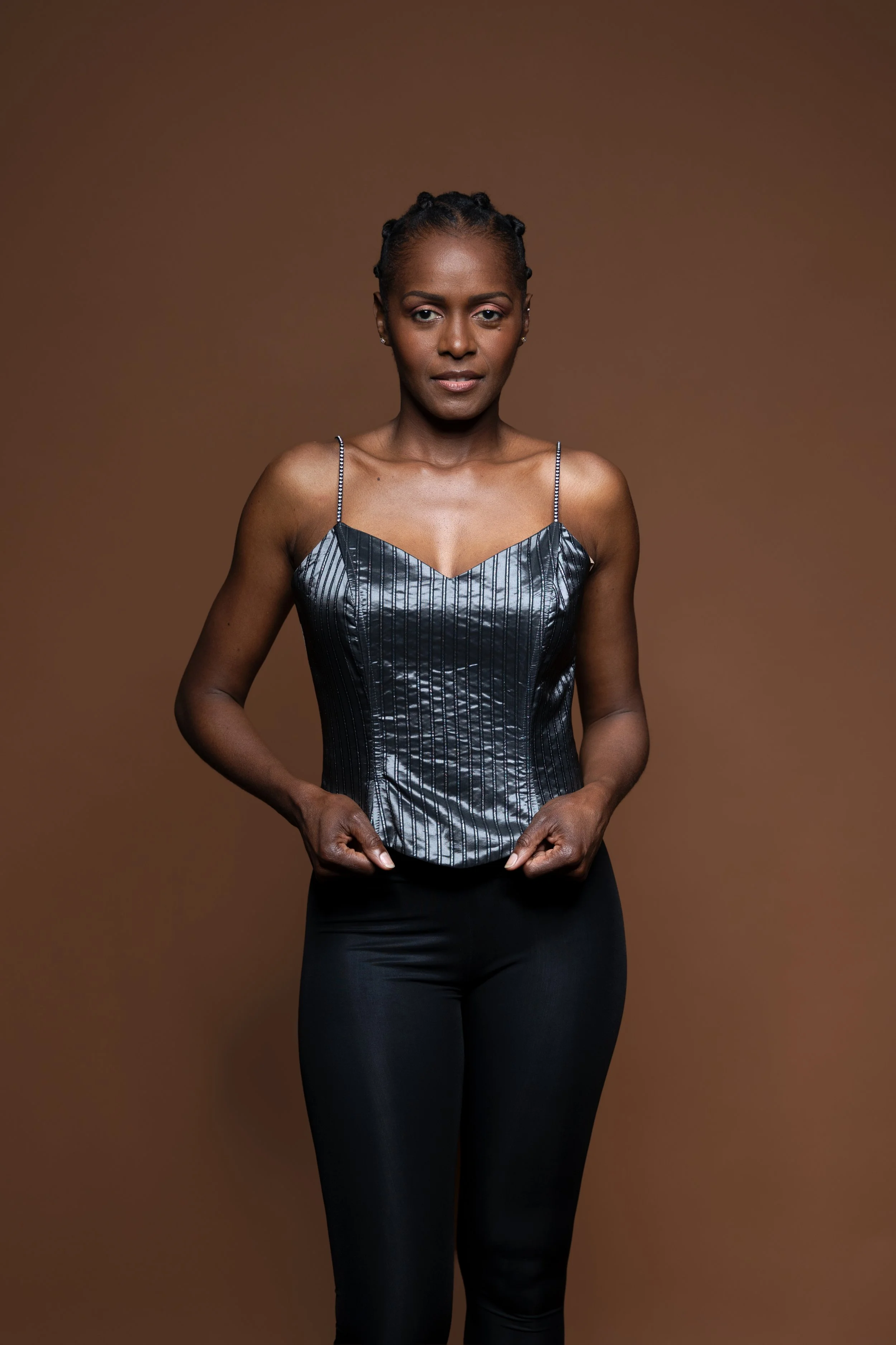 She faces the camera in a silver corset, holding its hem against a warm brown backdrop.