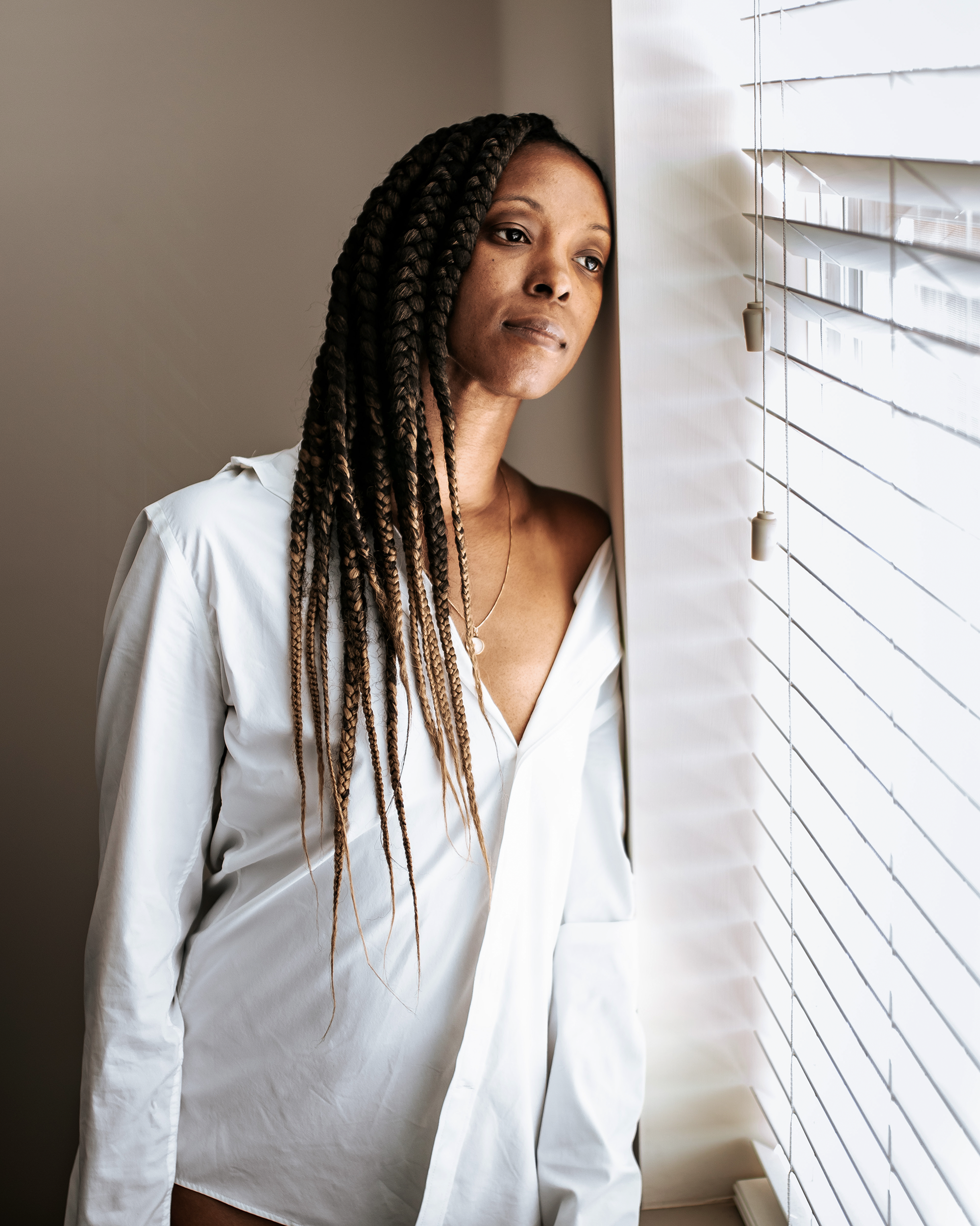 She pauses by the window, her gaze resting beyond the room. Light moves across the white shirt and pendant, leaving space for a moment of stillness.