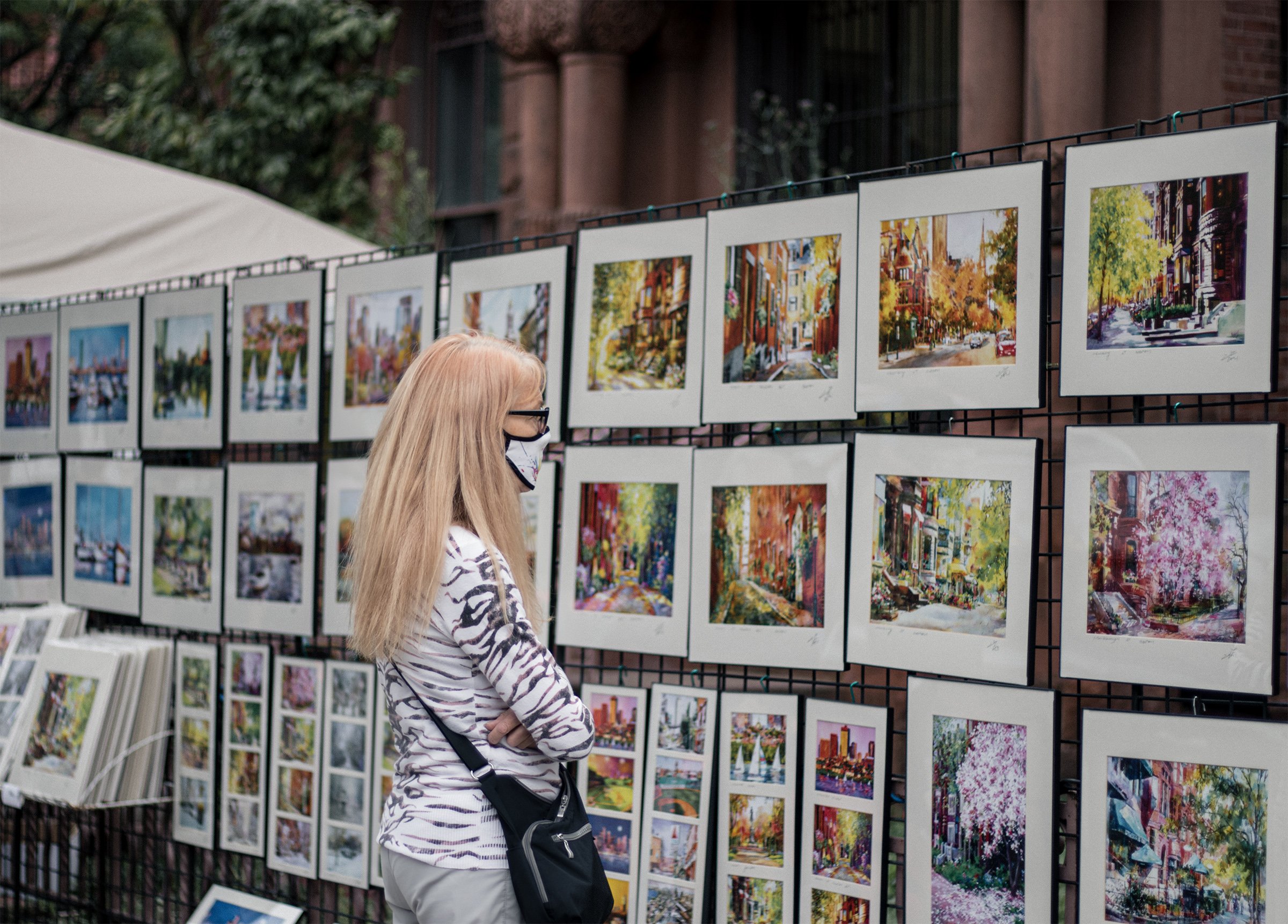 A woman with folded arms studies framed artwork at an outdoor display.