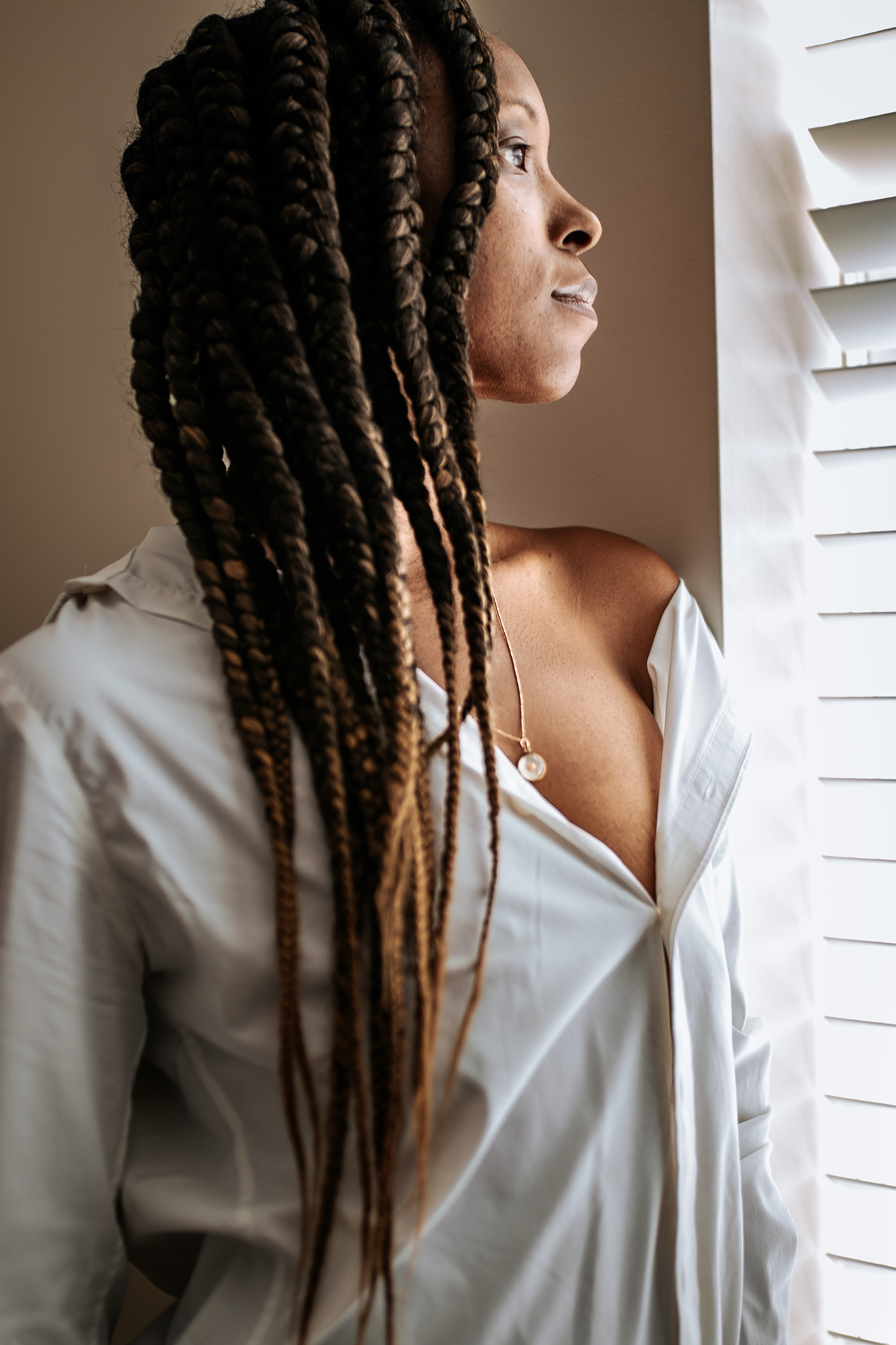 Beside the window, she turns toward the light. The quiet profile, loose white shirt, and soft fall of her braids give the portrait an intimate ease.