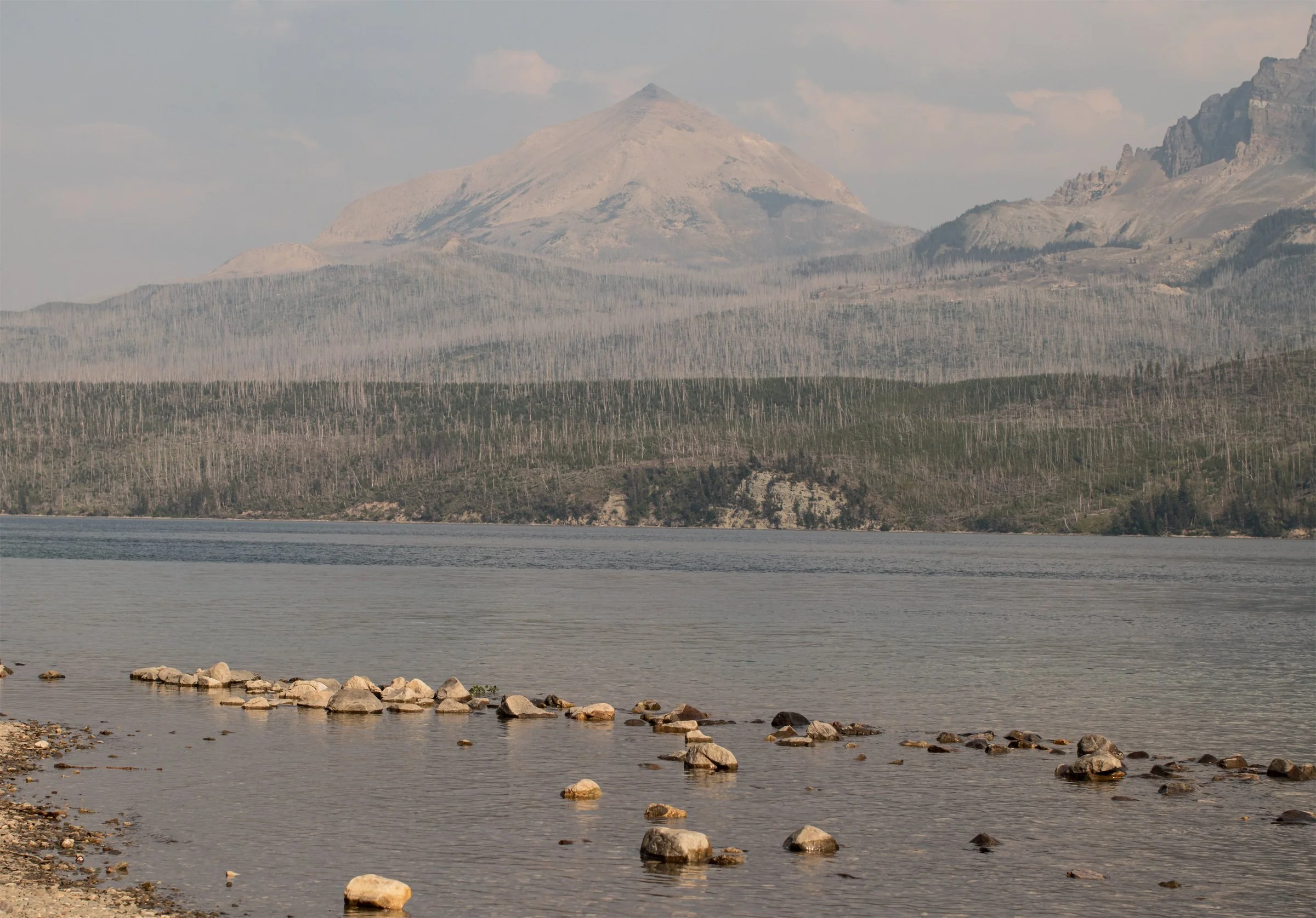 Clear lake water reveals stones below the surface, with mountains gathering beyond the shore. A quiet held at the waterline.
