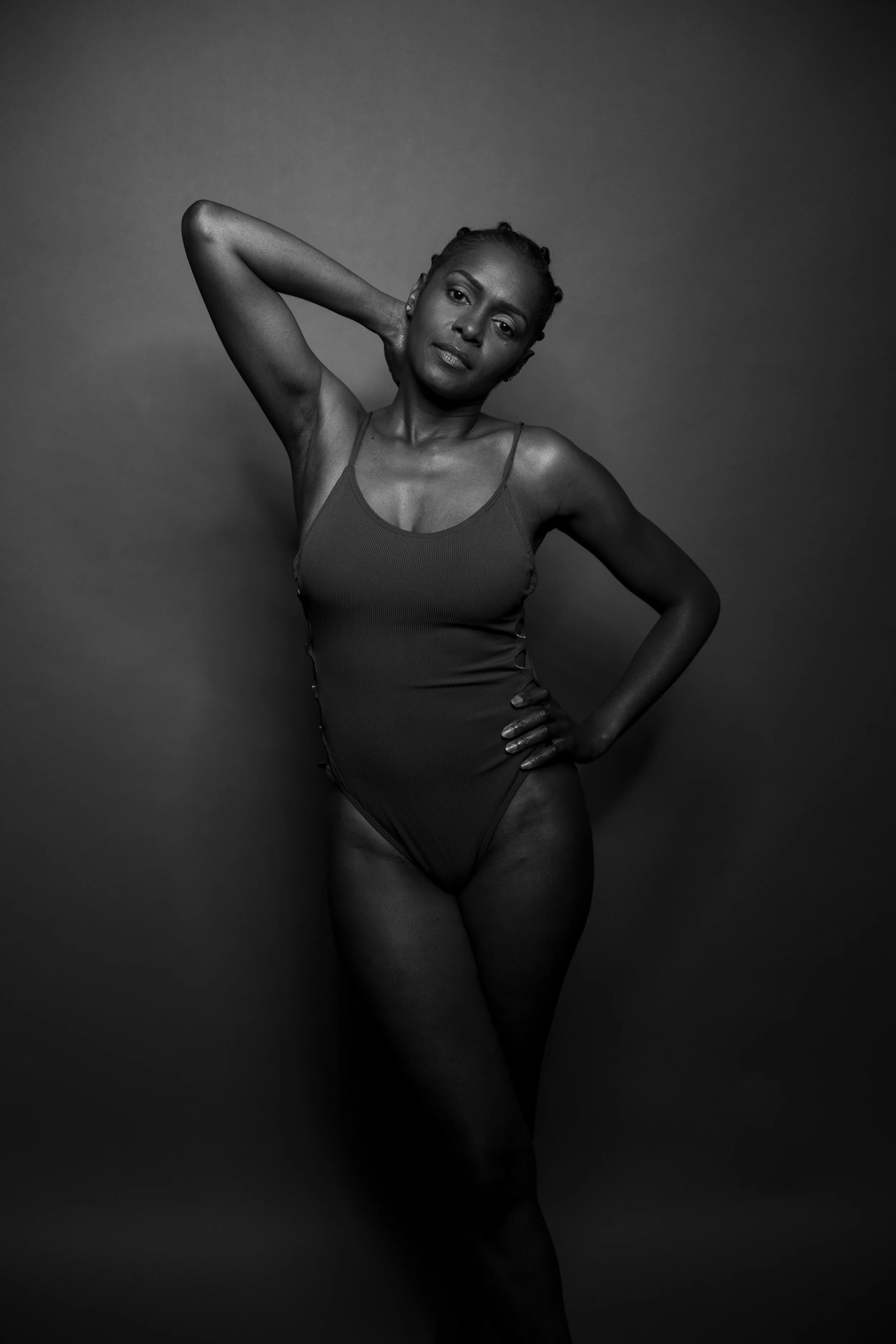 One arm curves behind her head, the other hand rests at her hip. Monochrome light traces the strength and grace of her silhouette.