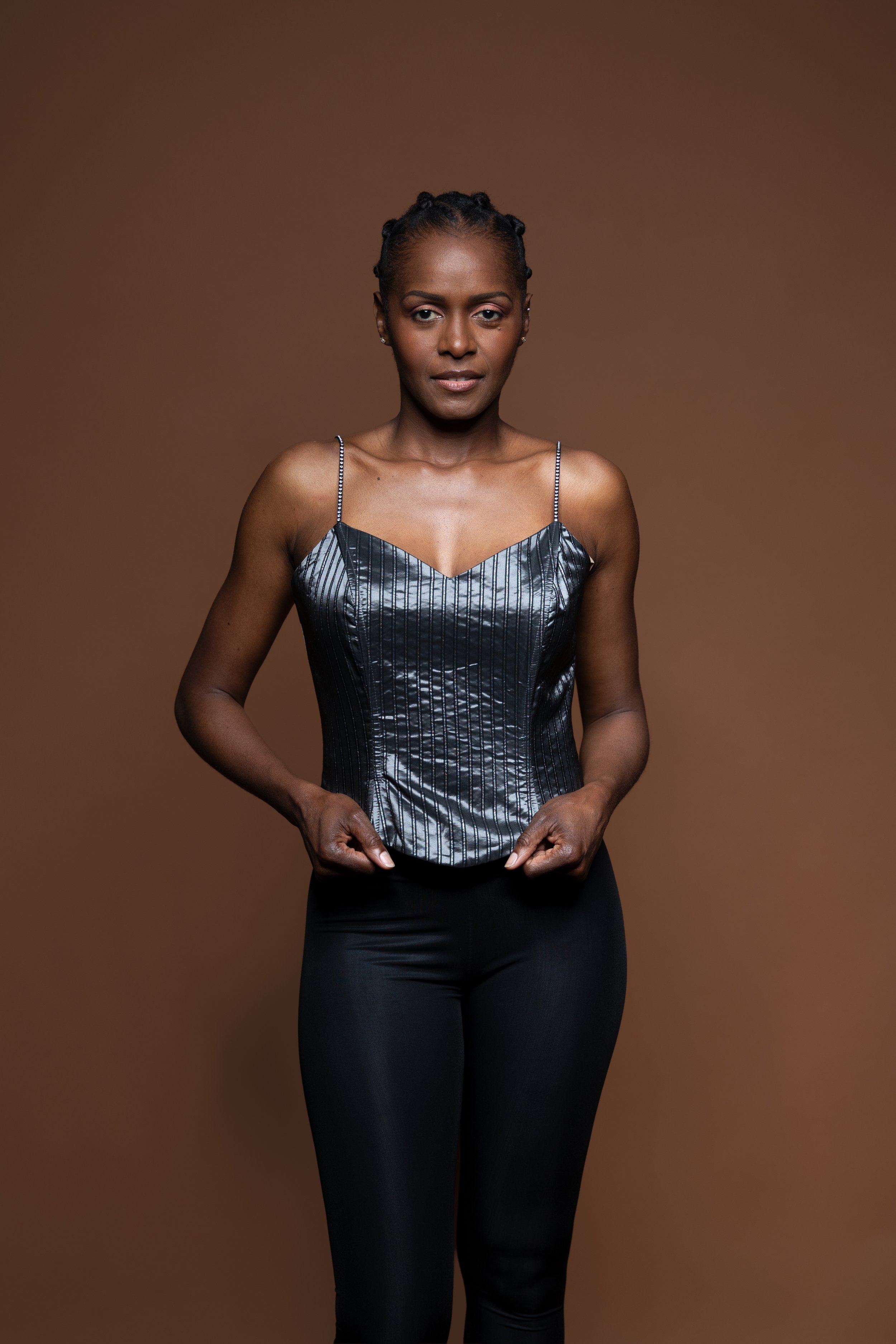 Silver catches the light as she holds the hem of her corset. Against warm brown, her direct expression gives the portrait its strength.