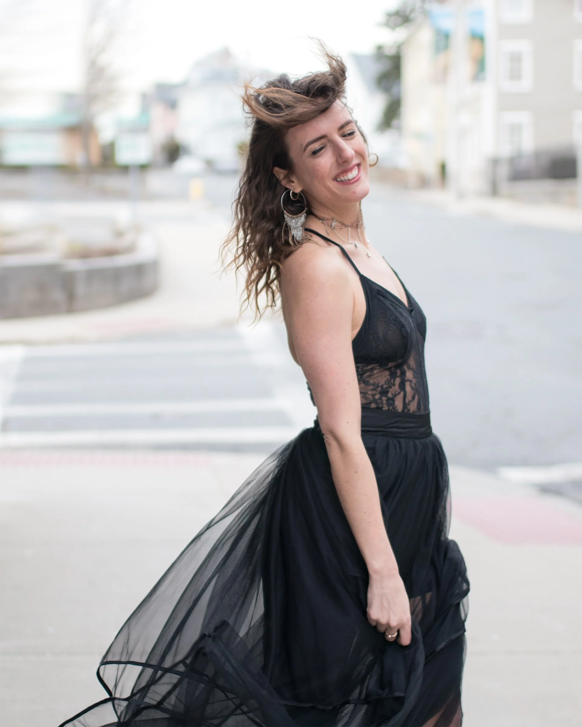 A woman in black lace smiles as wind lifts her hair against the city street.