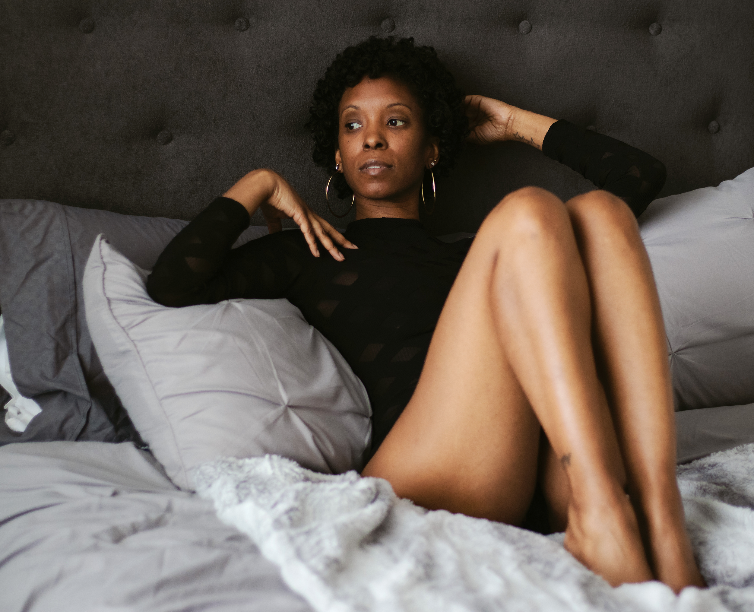 Reclining against gray pillows, she looks beyond the frame. Warm light and relaxed hands hold an unhurried moment.