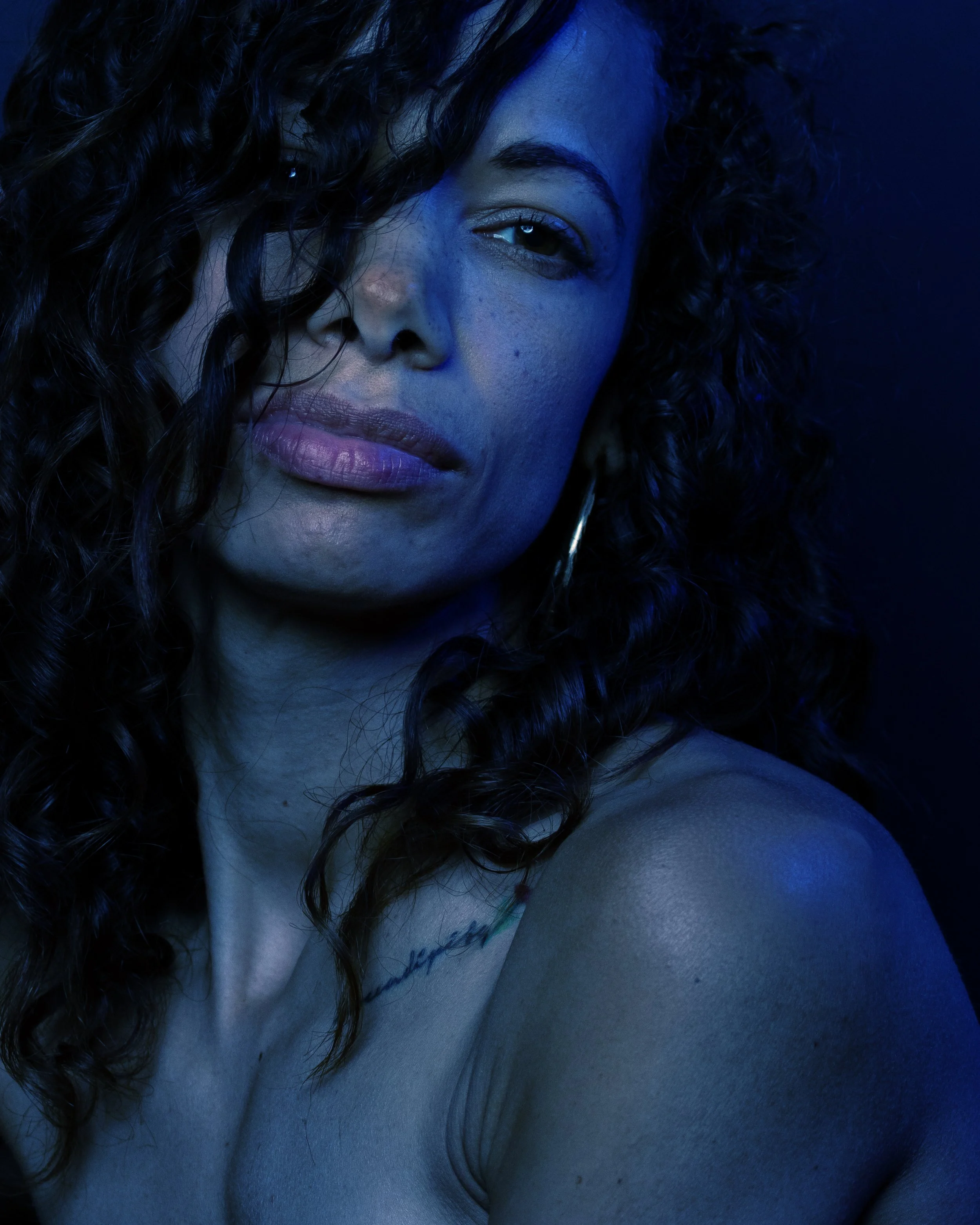 She looks into the camera through loose curls, her face and shoulder softly lit in blue.