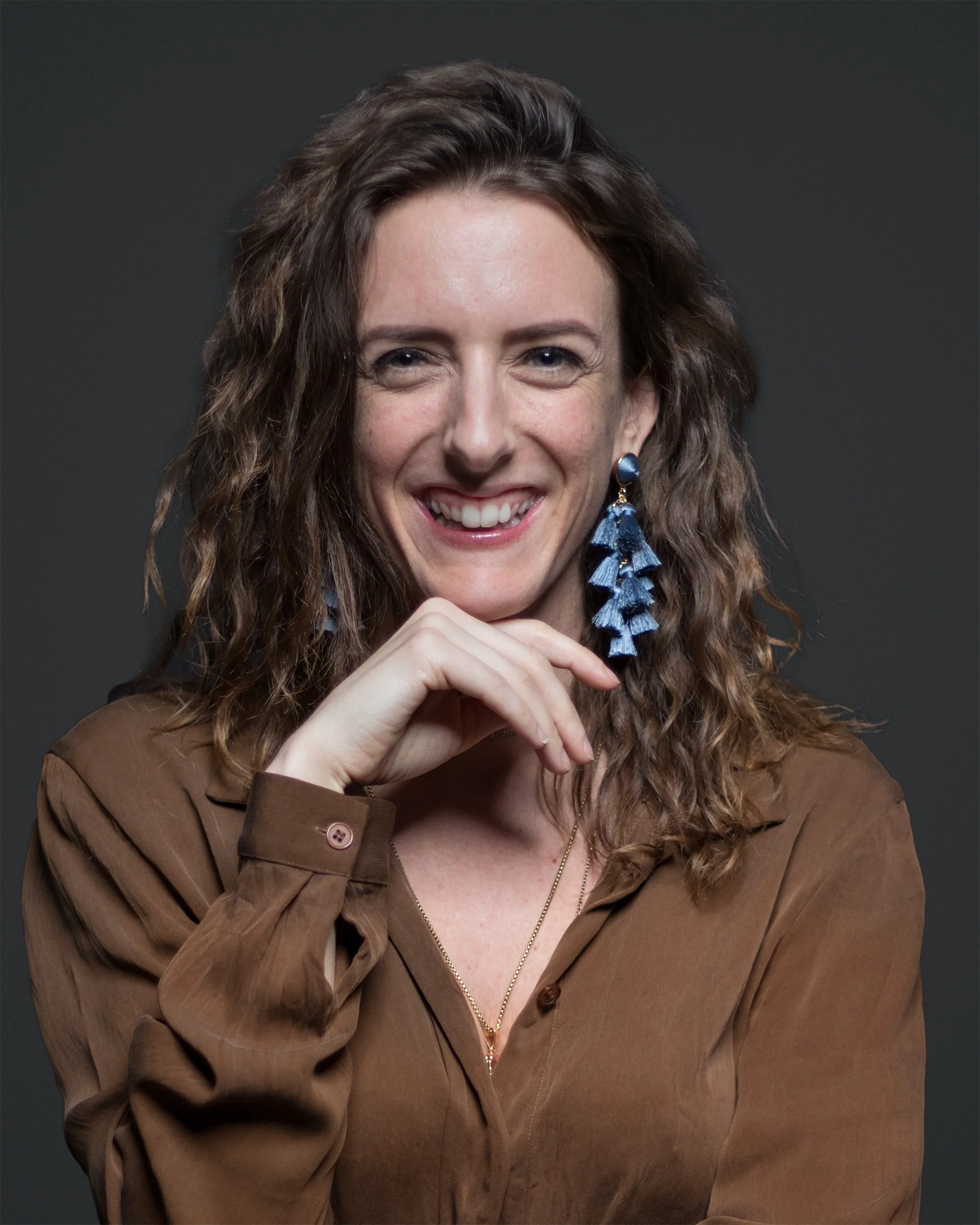 A smile meets the camera above a softly resting hand. Copper fabric and blue earrings bring warmth and color to an open, unguarded portrait.