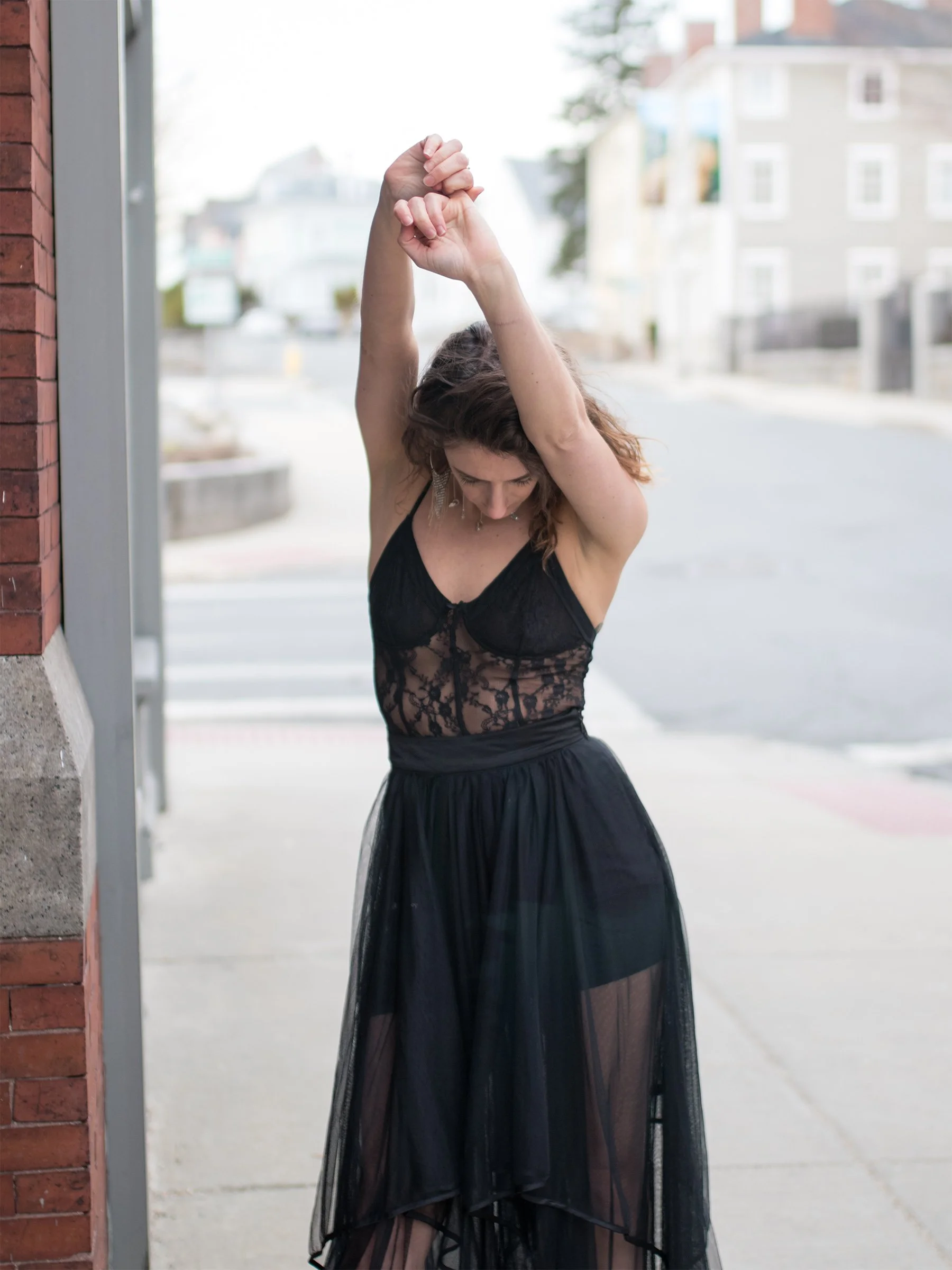 A woman in black lace lowers her head and raises her arms, framed by soft city light.