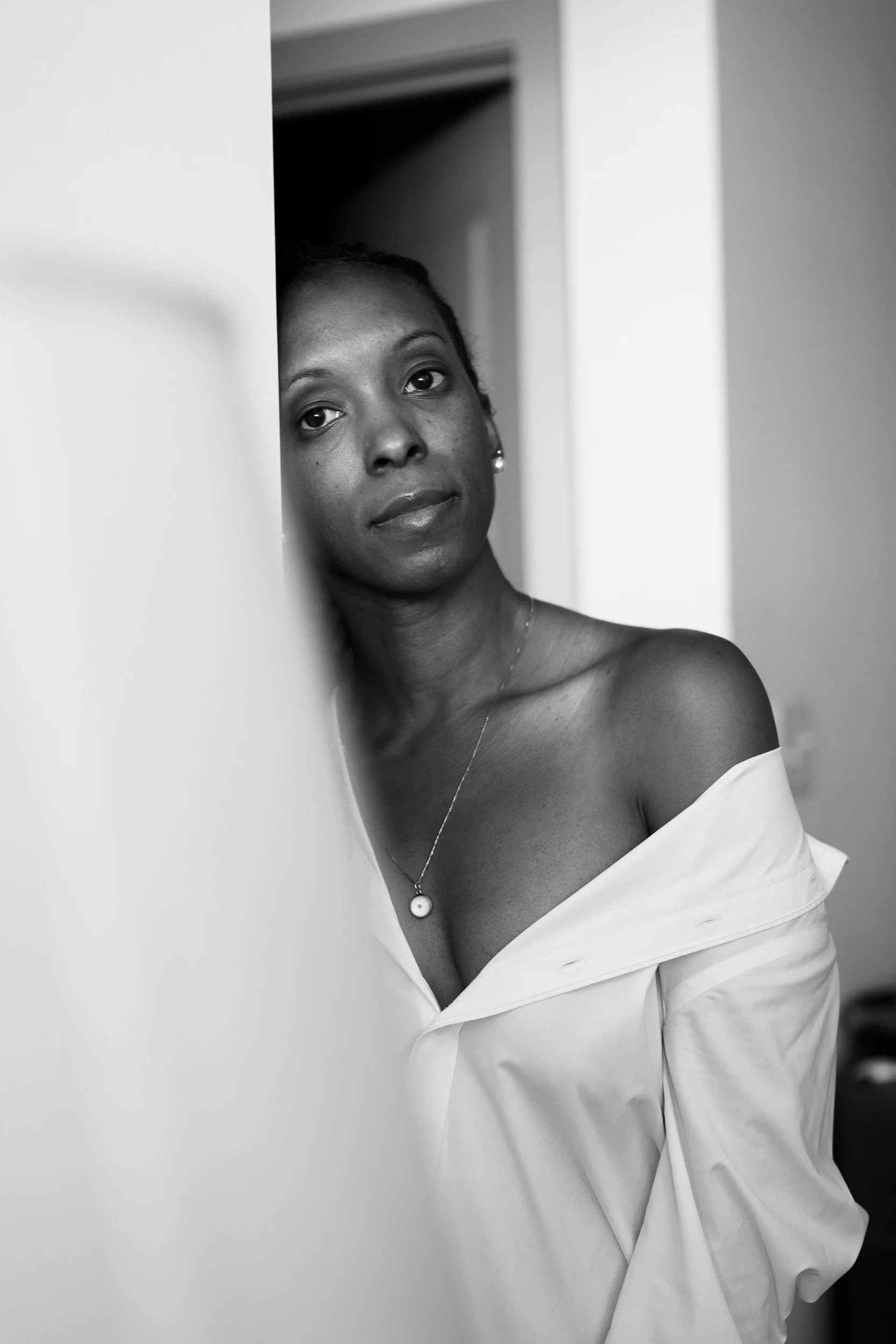 Framed by a softly blurred doorway, she meets the camera with a direct gaze. A white shirt and a delicate pendant bring quiet elegance to this intimate monochrome portrait.