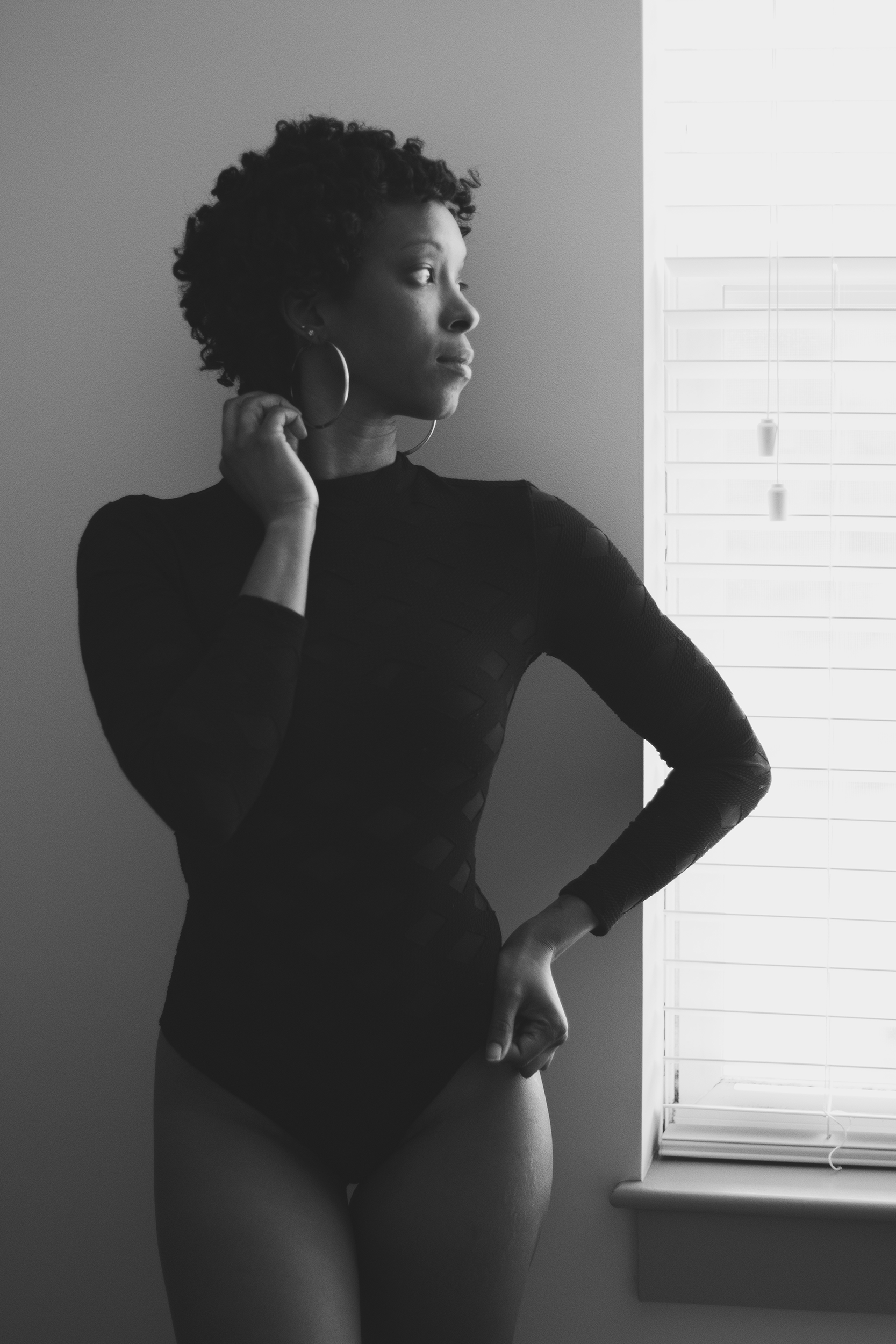 Standing beside the window, she looks toward the light, one hand resting near her earring. The curve of her arm and the depth of the shadows give this monochrome portrait a quiet strength.