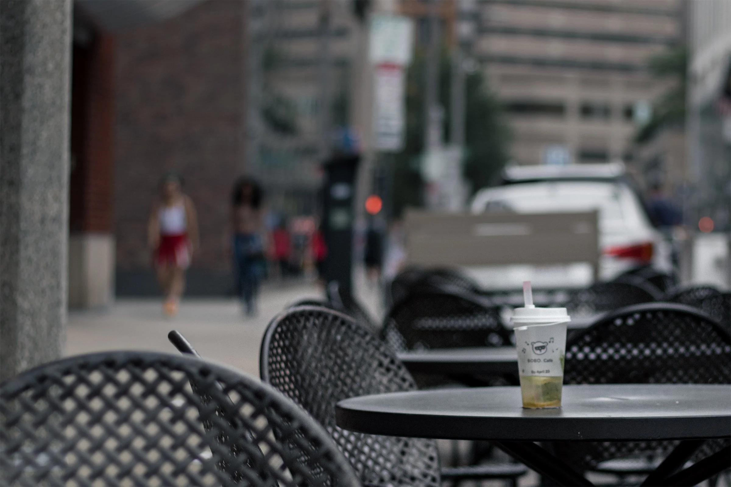 A single drink rests on a café table, surrounded by softly blurred chairs and street life.