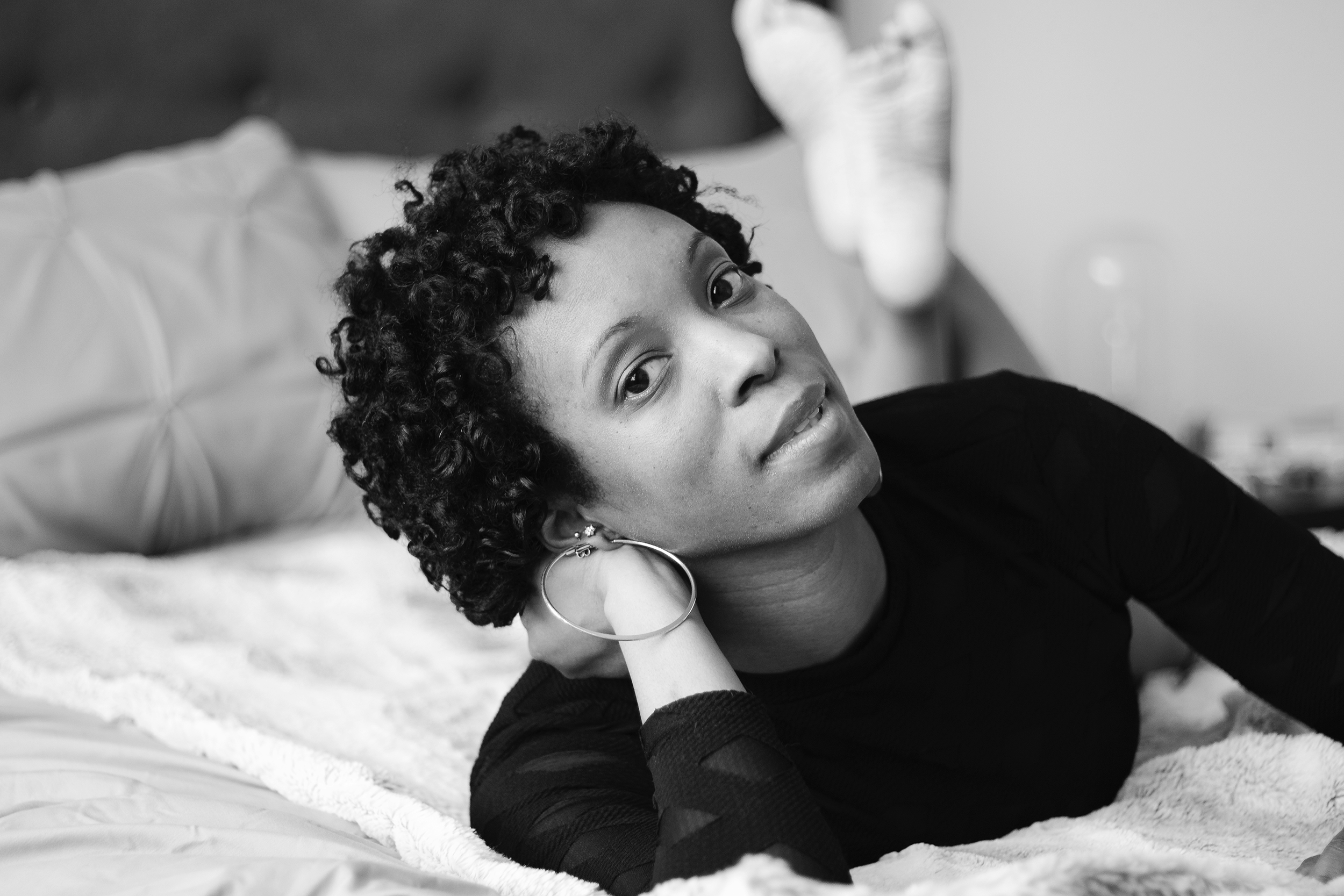 Resting her head in her hand, she meets the camera from the softness of the bed. The monochrome portrait holds the closeness of her gaze and the ease of an unguarded moment.