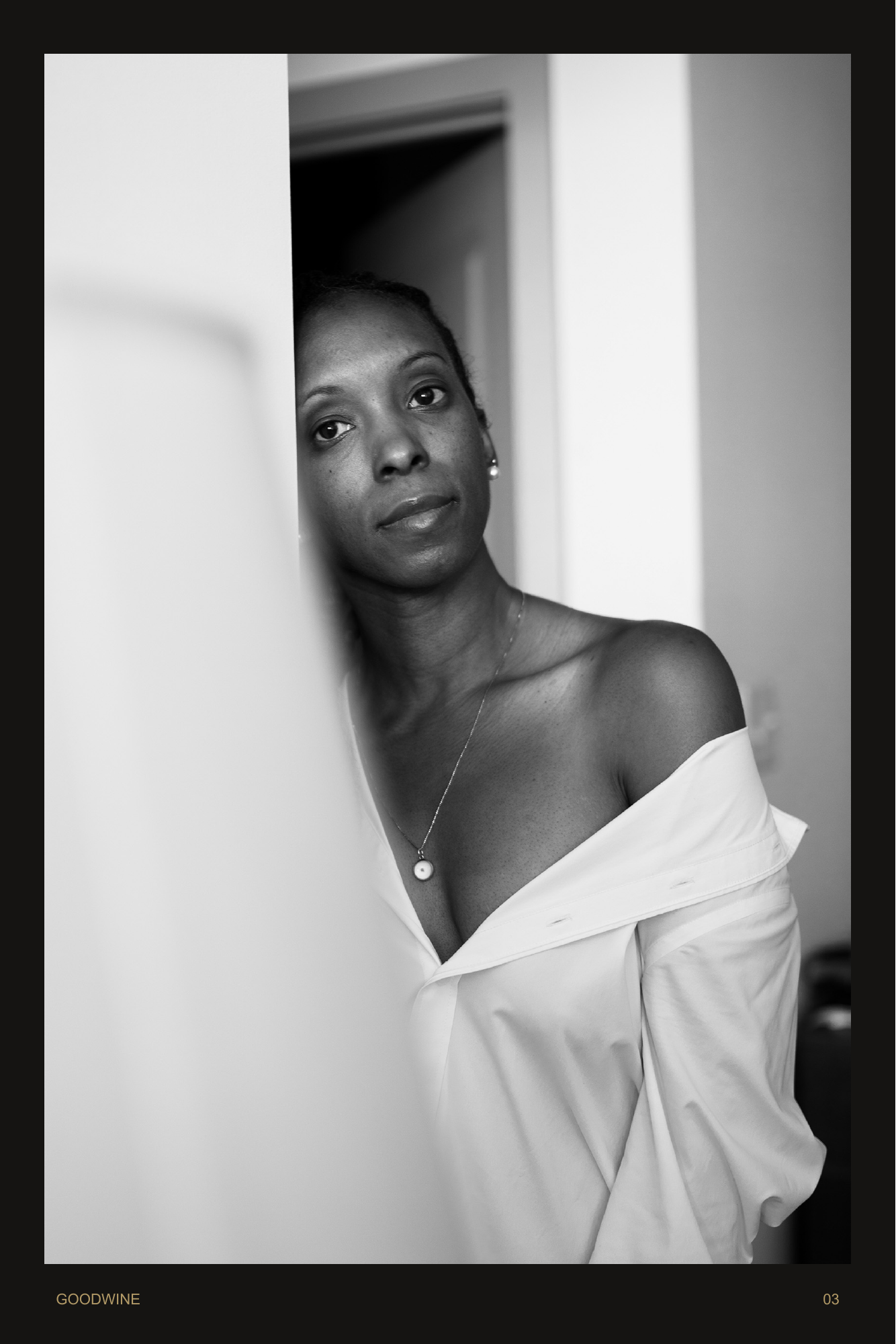 Page 3. She meets the camera from a doorway, wrapped in a loose white shirt. A quiet monochrome portrait.