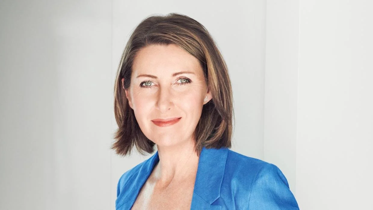Niki Bezzant wearing a blue blazer, smiling softly at the camera in front of a plain gray background.