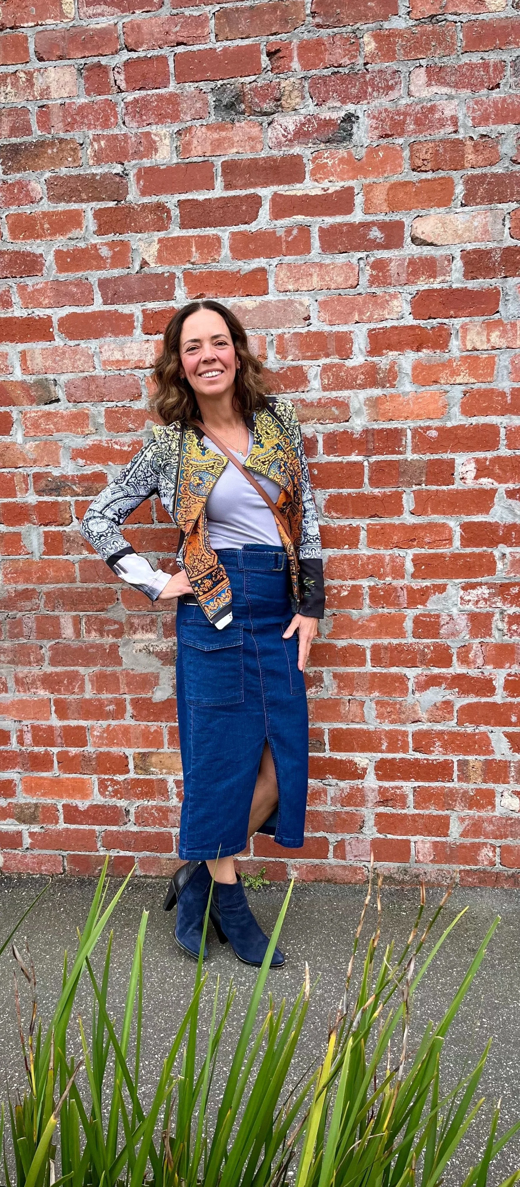 Woman smiling in front of brick wall, wearing patterned jacket, denim skirt, and blue boots.