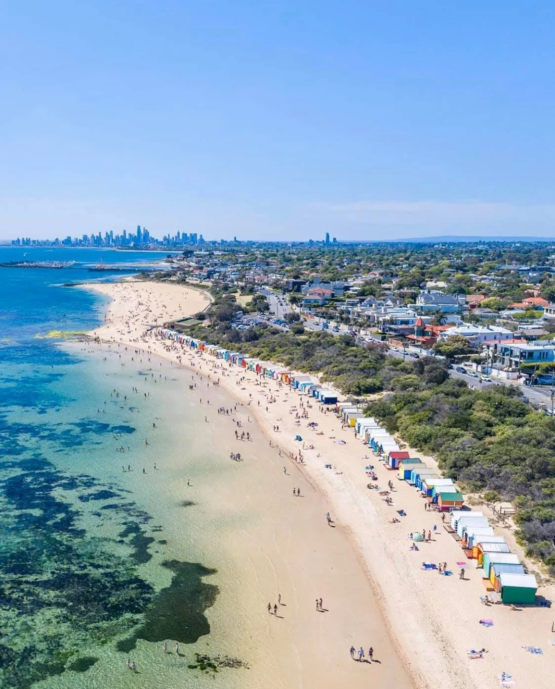 Aerial view of a beach with colorful bathing boxes along the shoreline, people enjoying the water, and a city skyline in the distance.