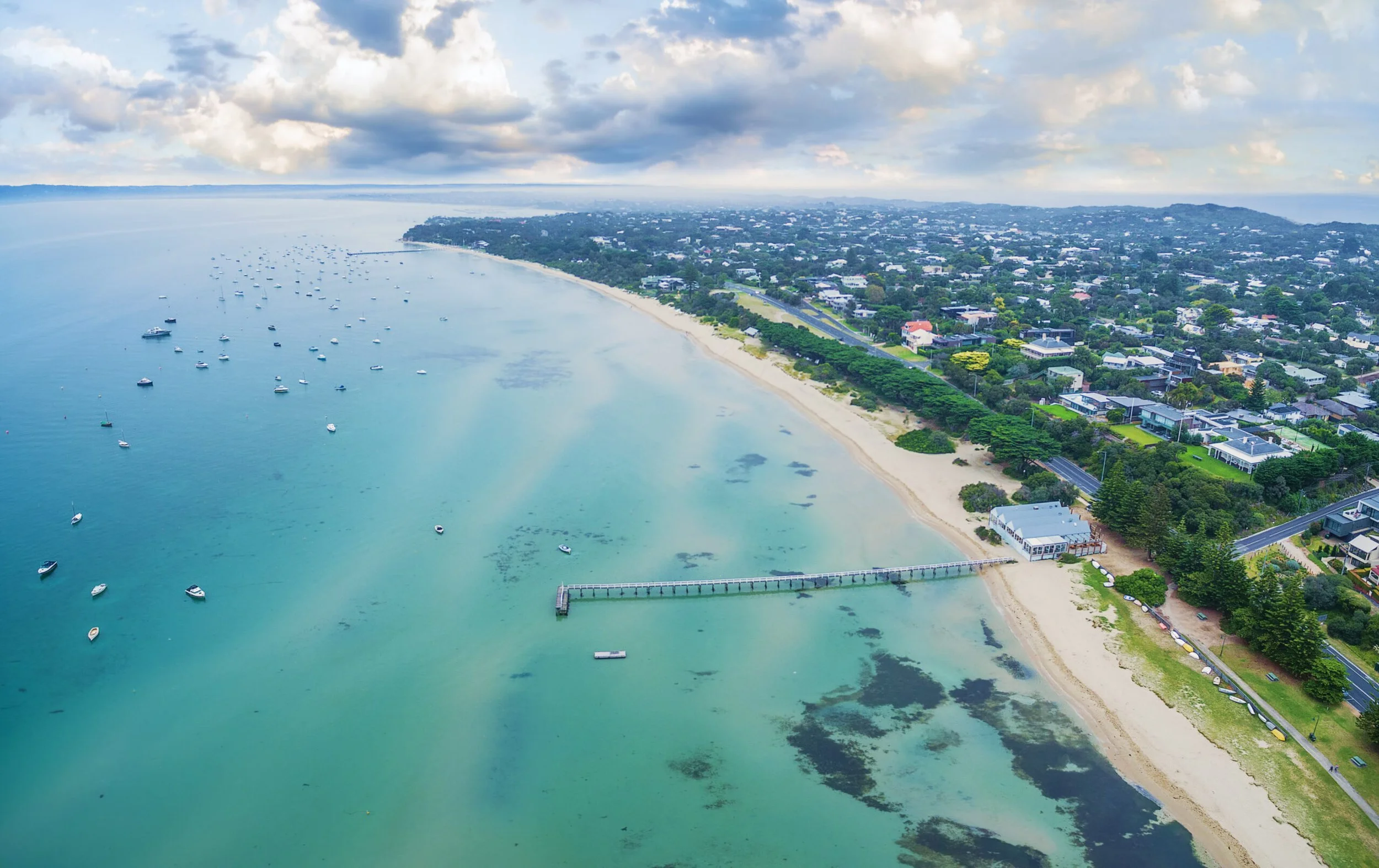 Aerial view of a coastal beach with a pier extending into the turquoise sea, numerous small boats anchored offshore, sandy shores, and lush greenery surrounding residential houses.