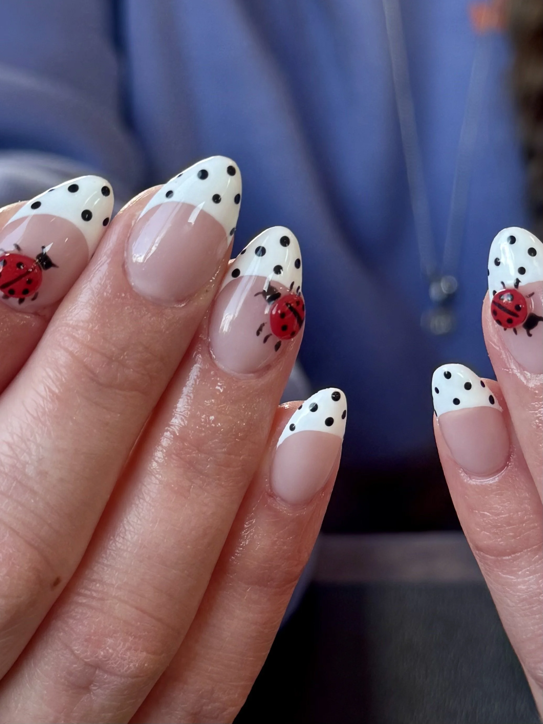 Close-up of a hand with decorated nails featuring white polish with black polka dots and a red ladybug design, set against a blurred dark blue background.