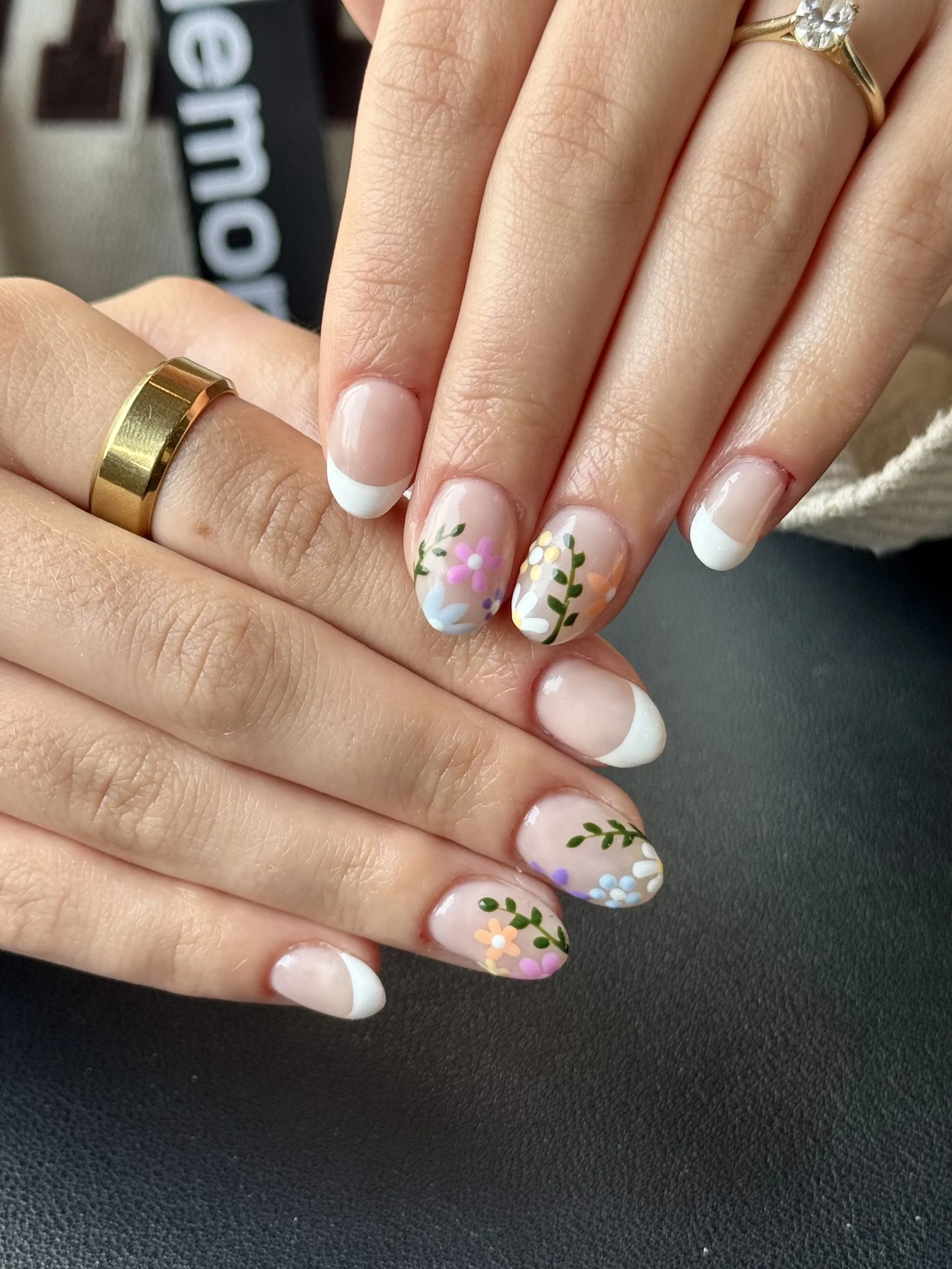 Close-up of a person's hand with a gold wedding band and a ring with a large clear gemstone. The nails are decorated with white French tips and floral designs in pastel colors.