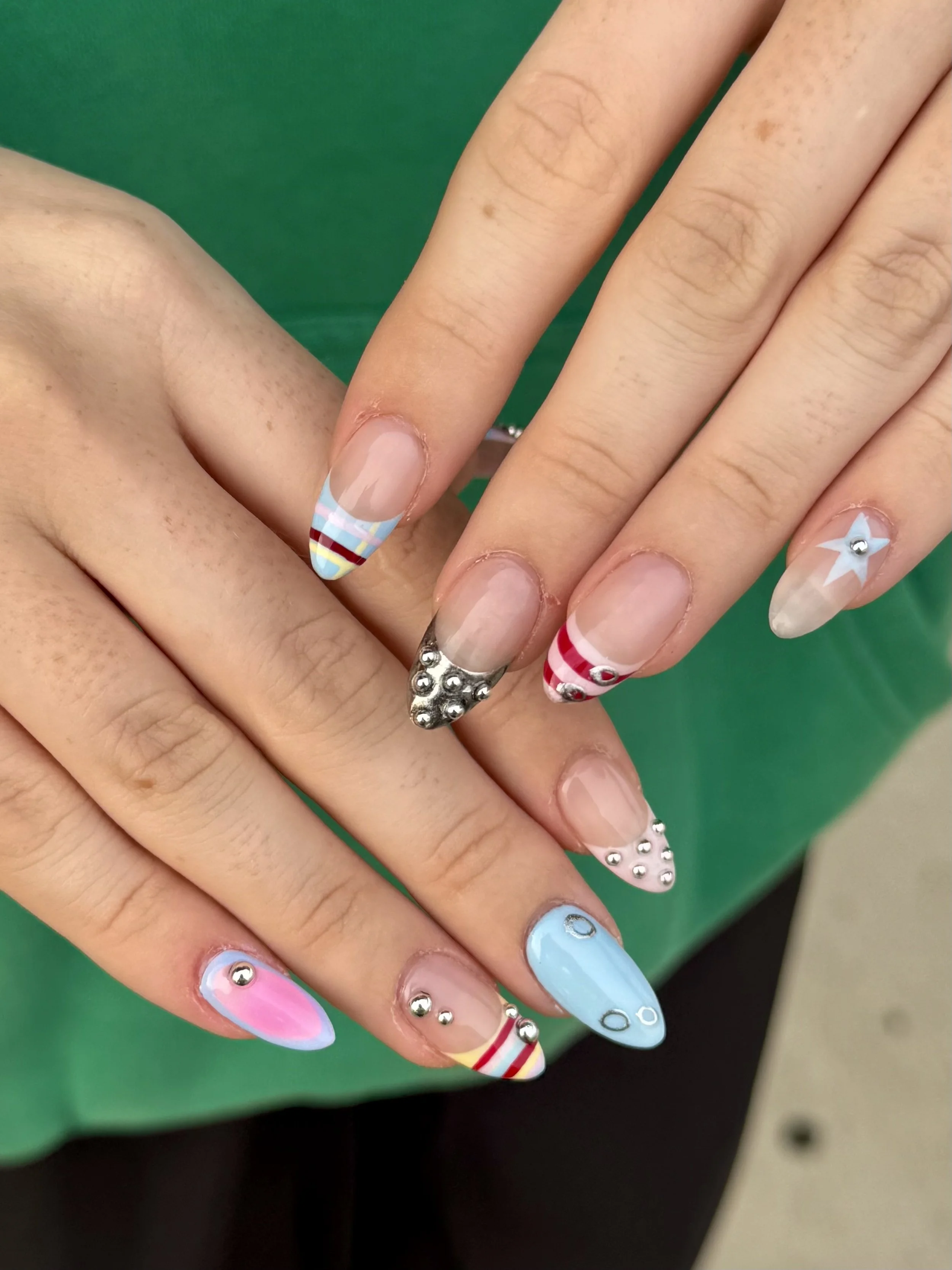 Close-up of hands with colorful, decorated nail art and silver embellishments, against a green background.