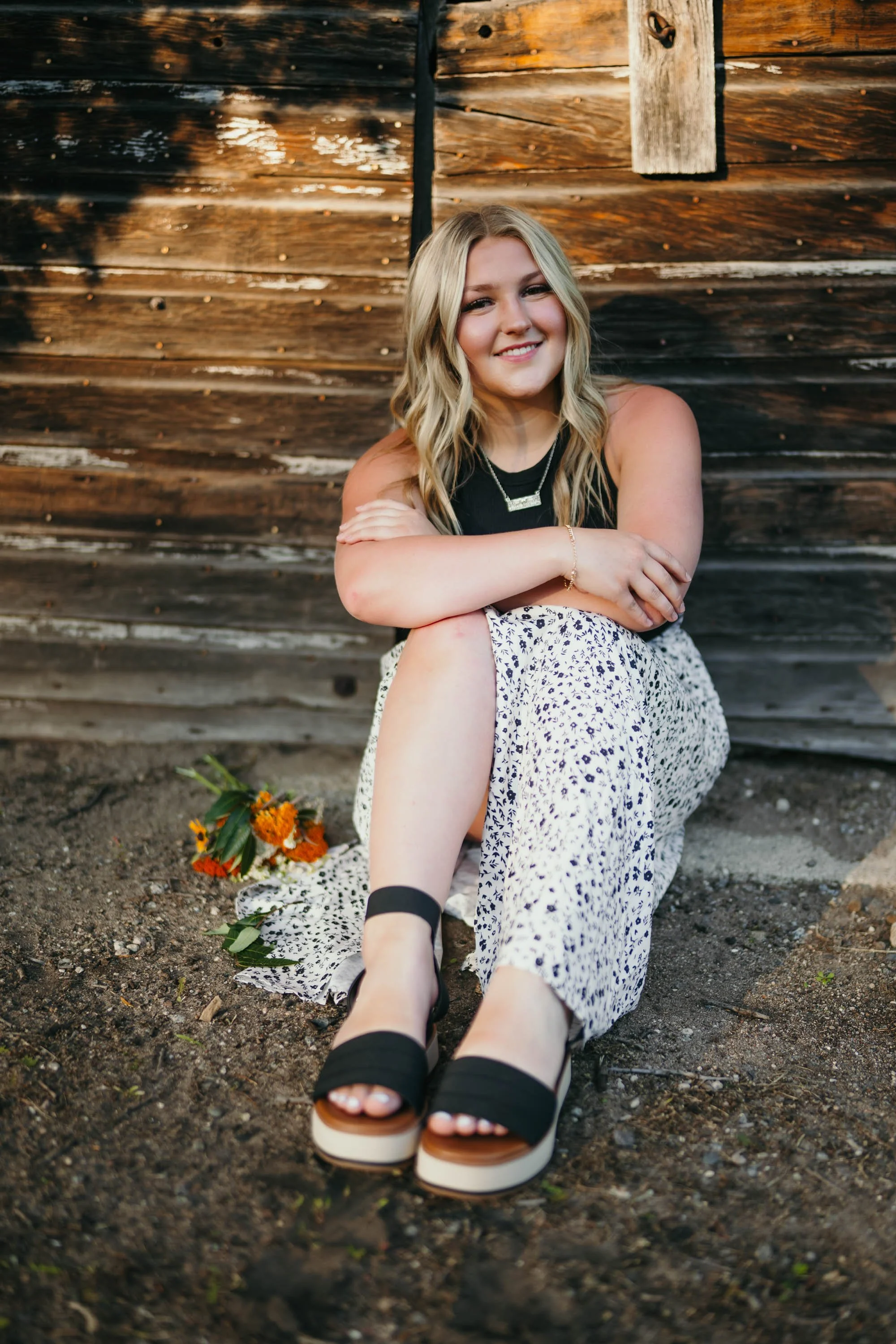 A young woman with blonde hair sitting on the ground in front of a wooden barn wall, smiling and resting her arms on her knees. She wears a black sleeveless top, a flowy printed skirt, black sandals, and jewelry. A bouquet of orange flowers lies on the ground beside her, and the photo is taken during sunset or golden hour.
