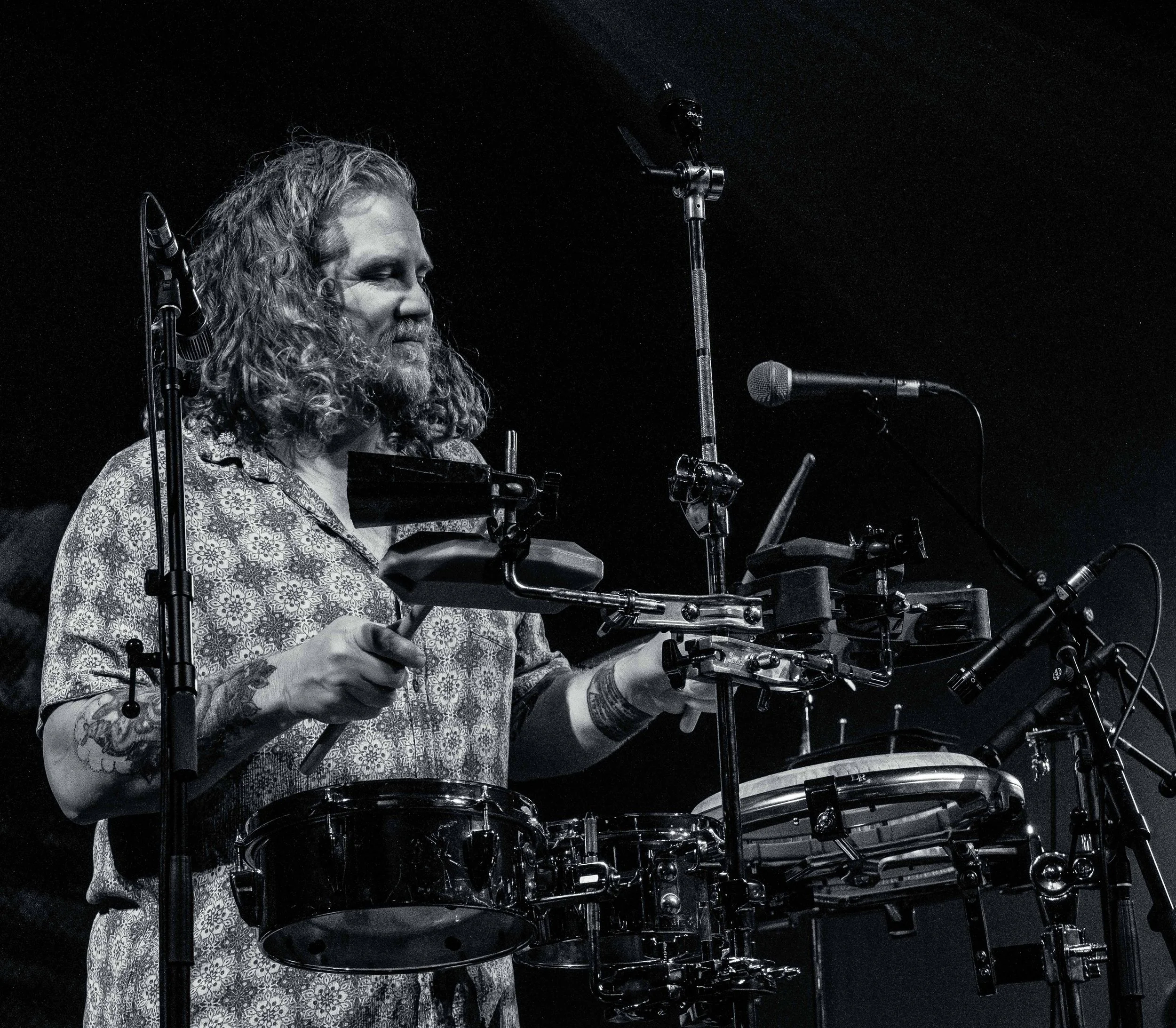 Black and white photo of a musician with long curly hair playing drums on stage, with microphones nearby.