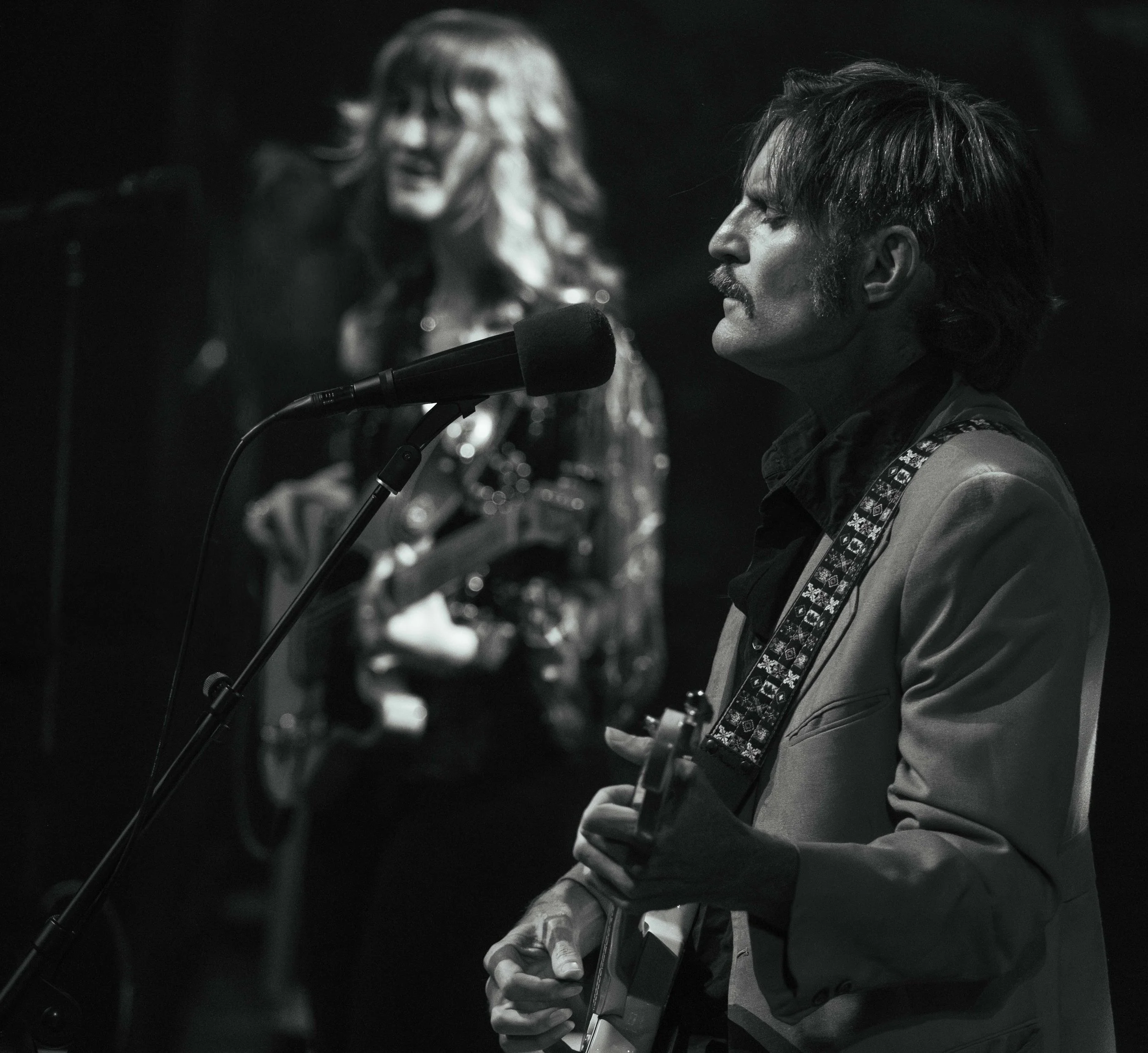 A black and white photo of a man playing an electric guitar and singing into a microphone, with a woman singing in the background.