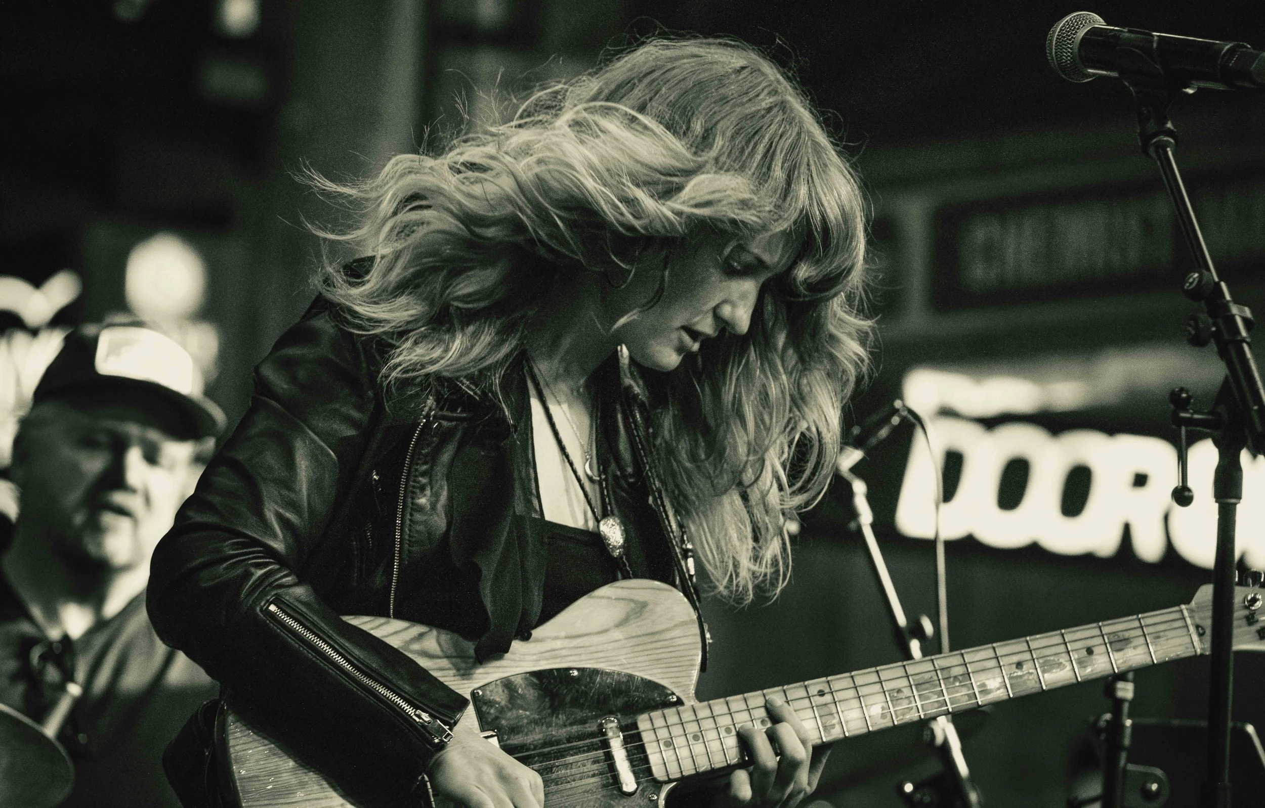 A woman with long wavy hair playing an acoustic guitar and singing into a microphone during a live performance, with a man in the background.