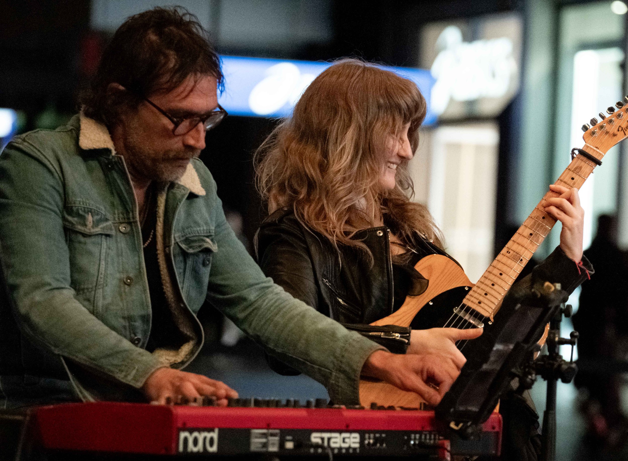 Two musicians playing instruments, one playing a keyboard and the other playing a guitar, in a dimly lit setting.