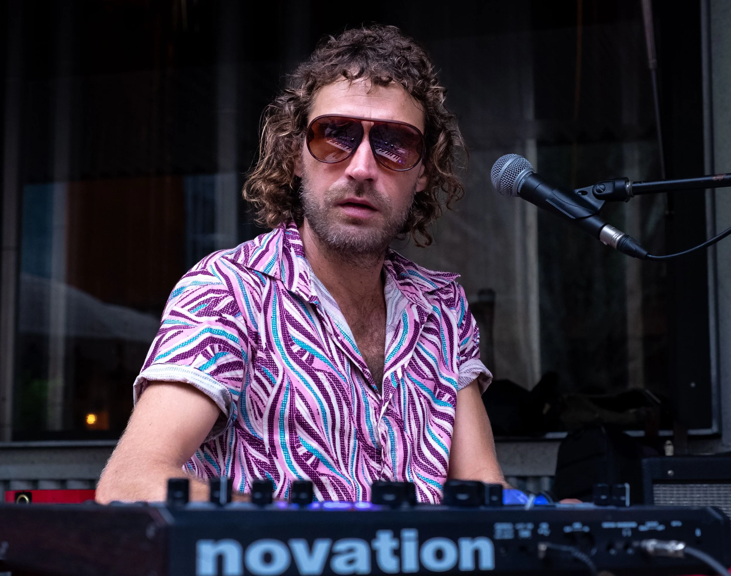 A man with curly hair wearing sunglasses and a colorful, patterned shirt, performing at a music event with a keyboard and a microphone.