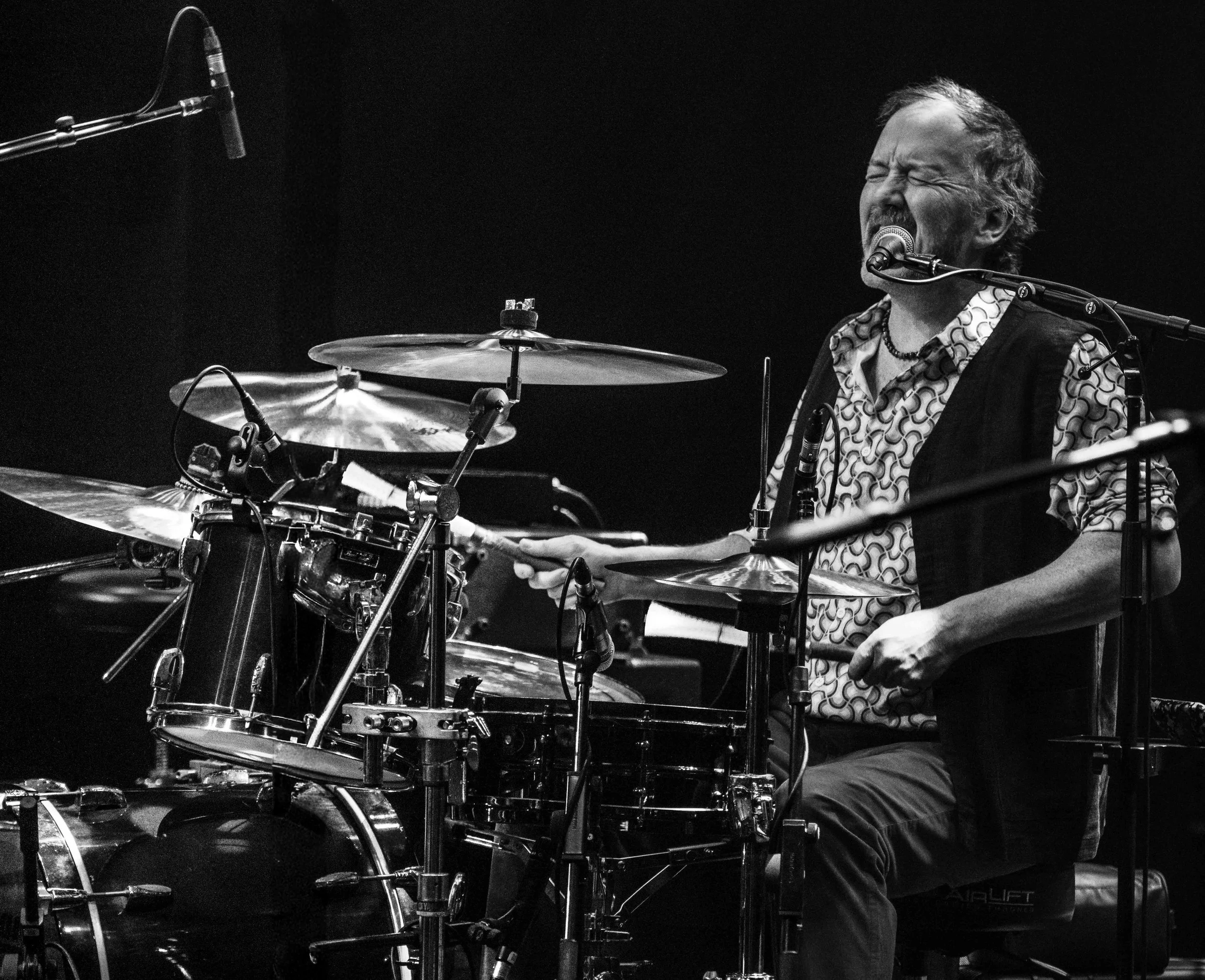Black and white image of a man playing drums passionately, with eyes closed and mouth open, captured mid-performance, microphone in front of him, dark background.