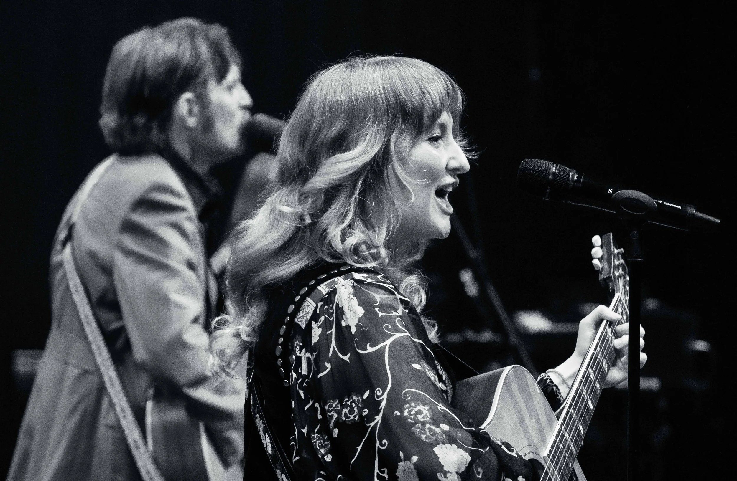 Two women performing music on stage: one singing into a microphone and playing an acoustic guitar, the other singing into a microphone, both in black and white.