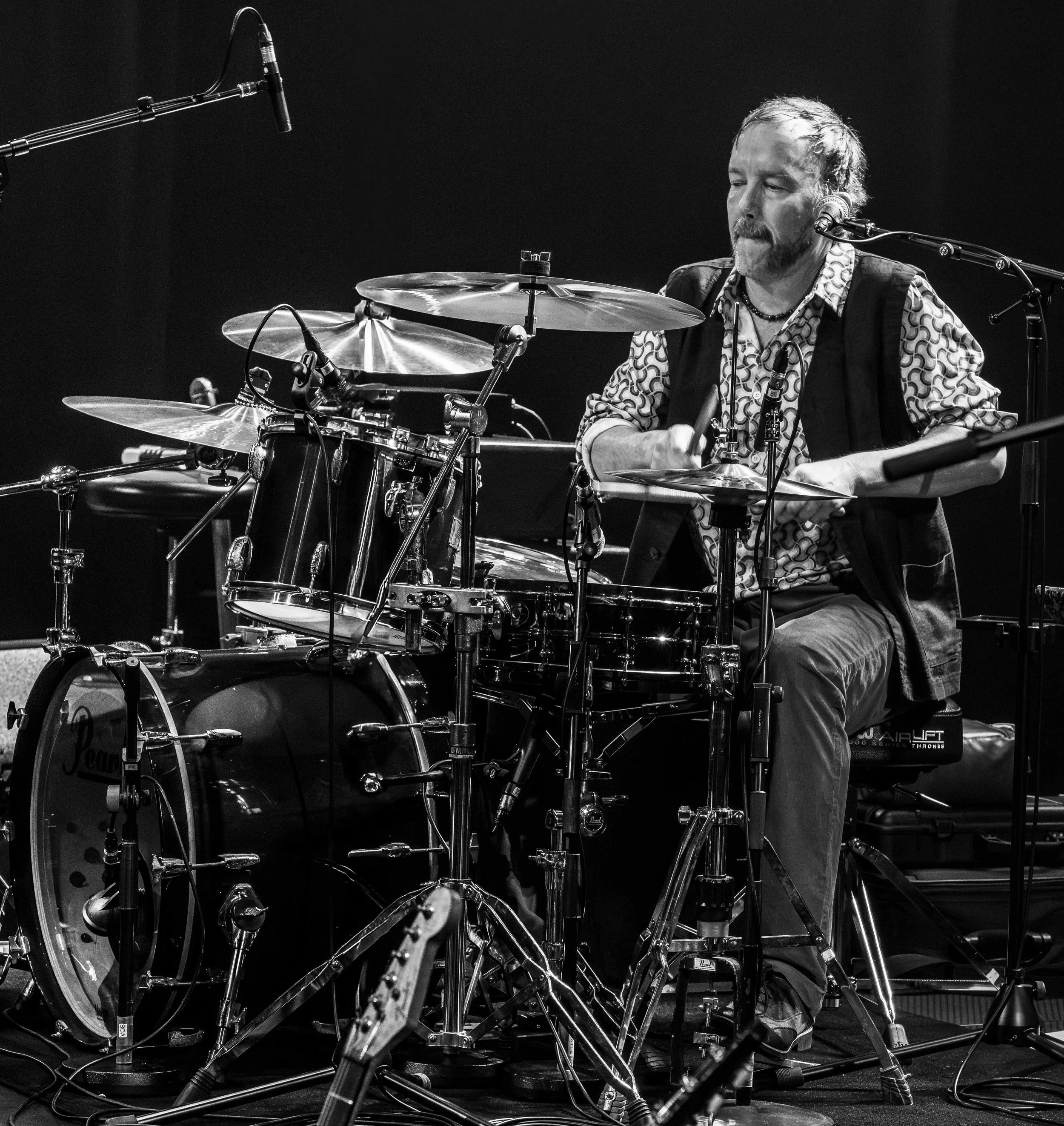 Musician playing drums on stage with microphones and equipment, in black and white.