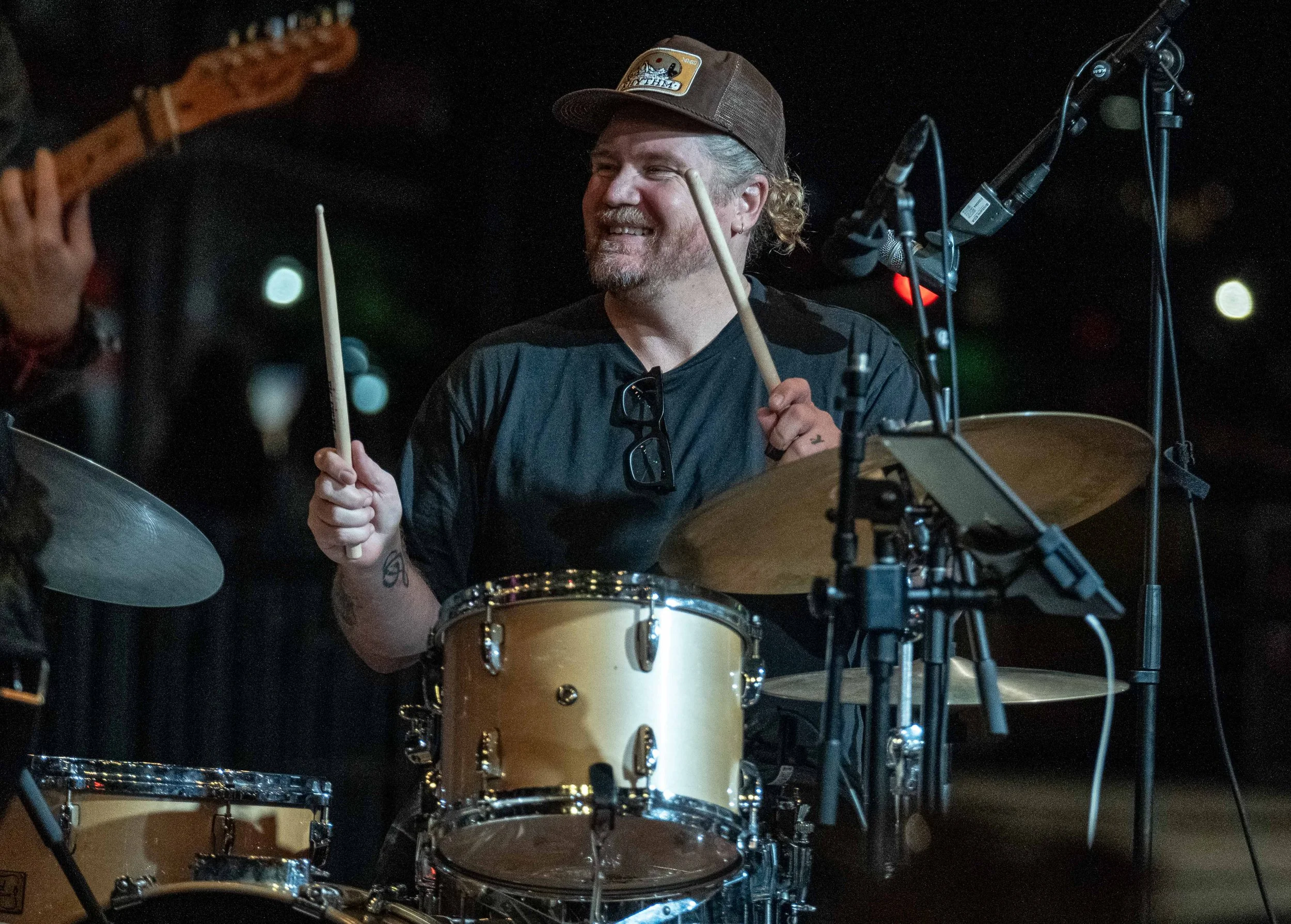 A man with a beard, wearing a brown cap and black shirt, smiling while playing drums. He is holding drumsticks and surrounded by microphone equipment in a dark setting.