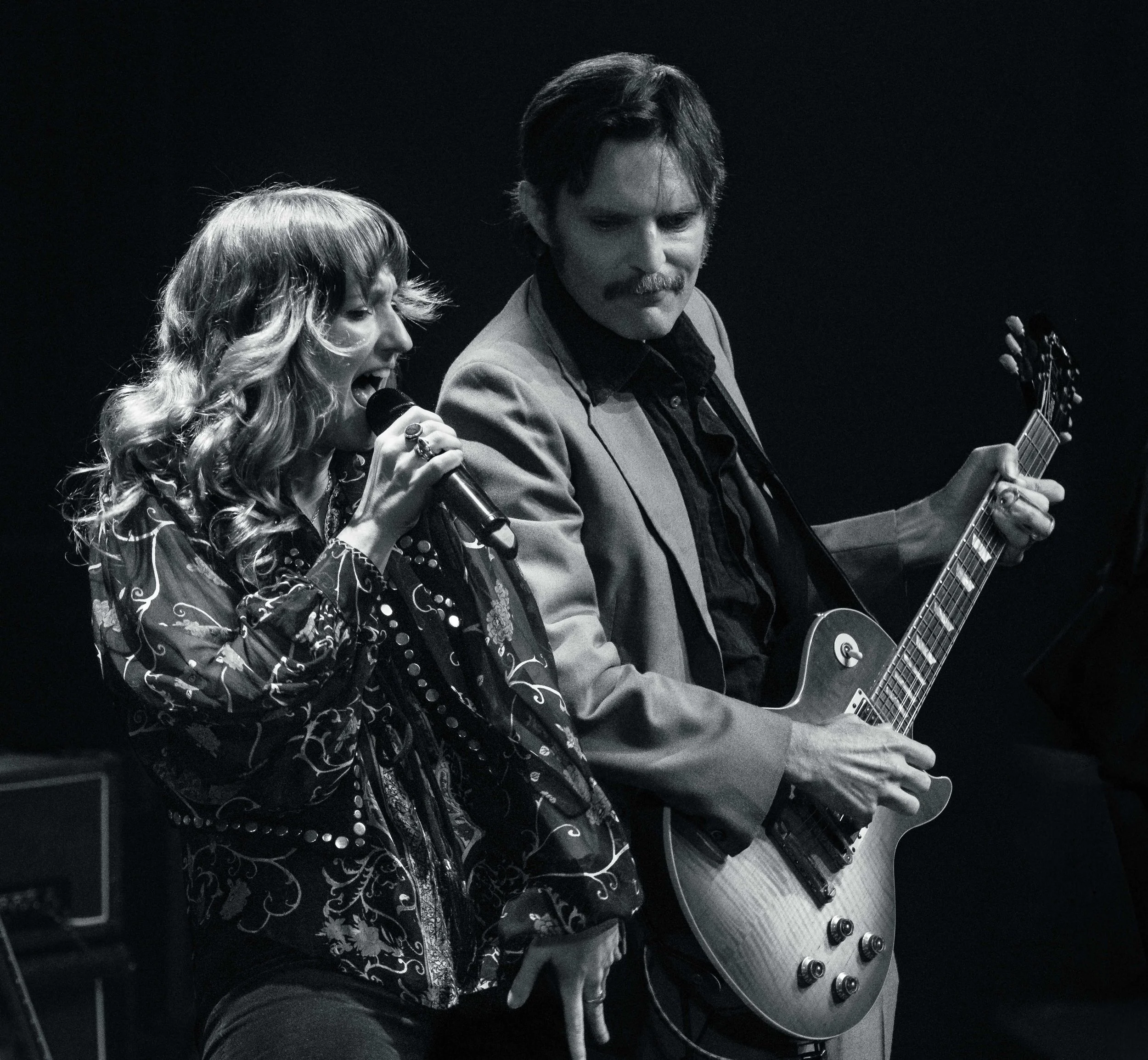 A black and white photo of a woman singing into a microphone and a man playing an acoustic guitar on stage.