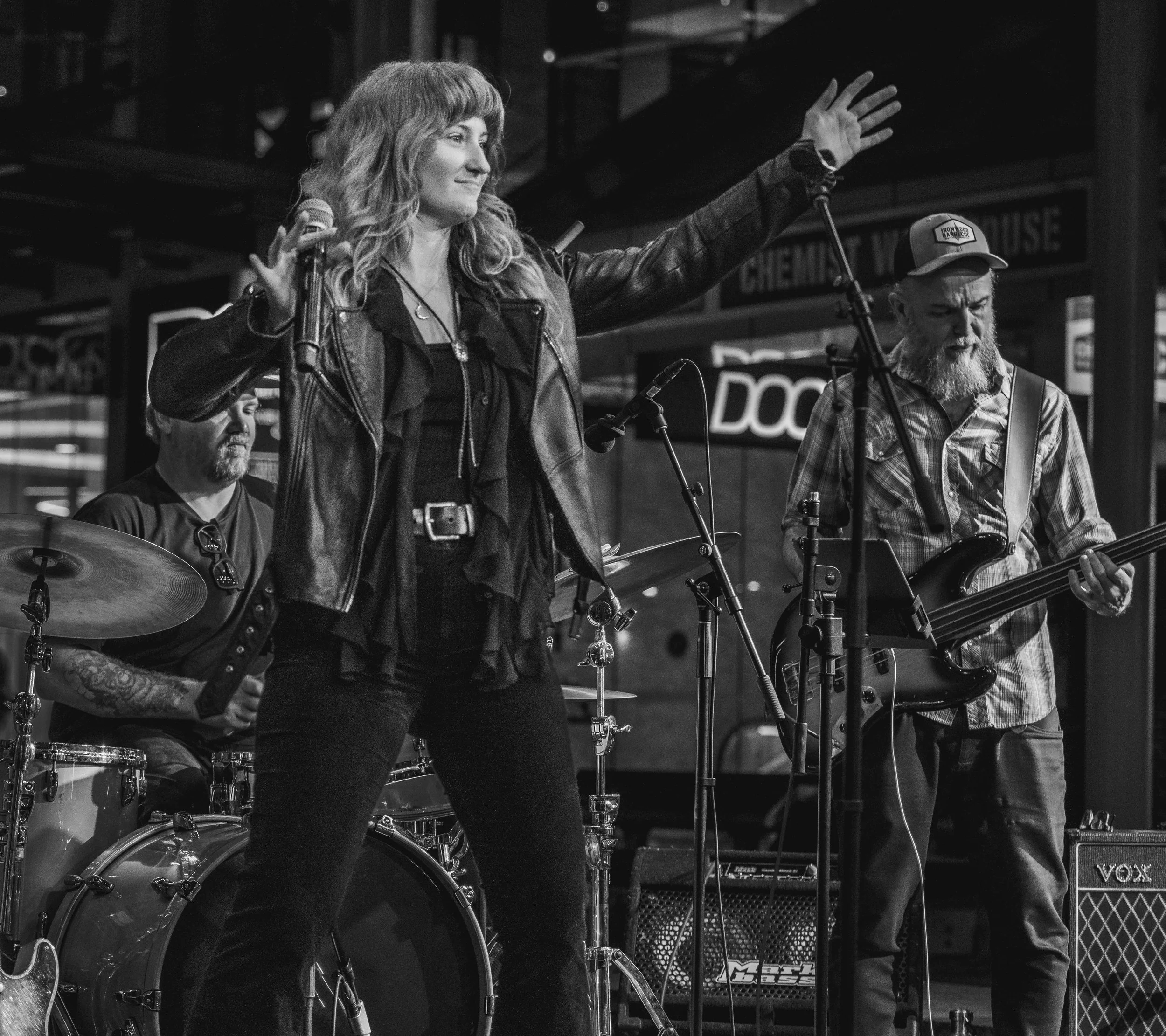 A woman with curly hair and tattoos on her arm singing into a microphone on stage, accompanied by a man with a beard wearing a cap and playing a guitar, during a live music performance.