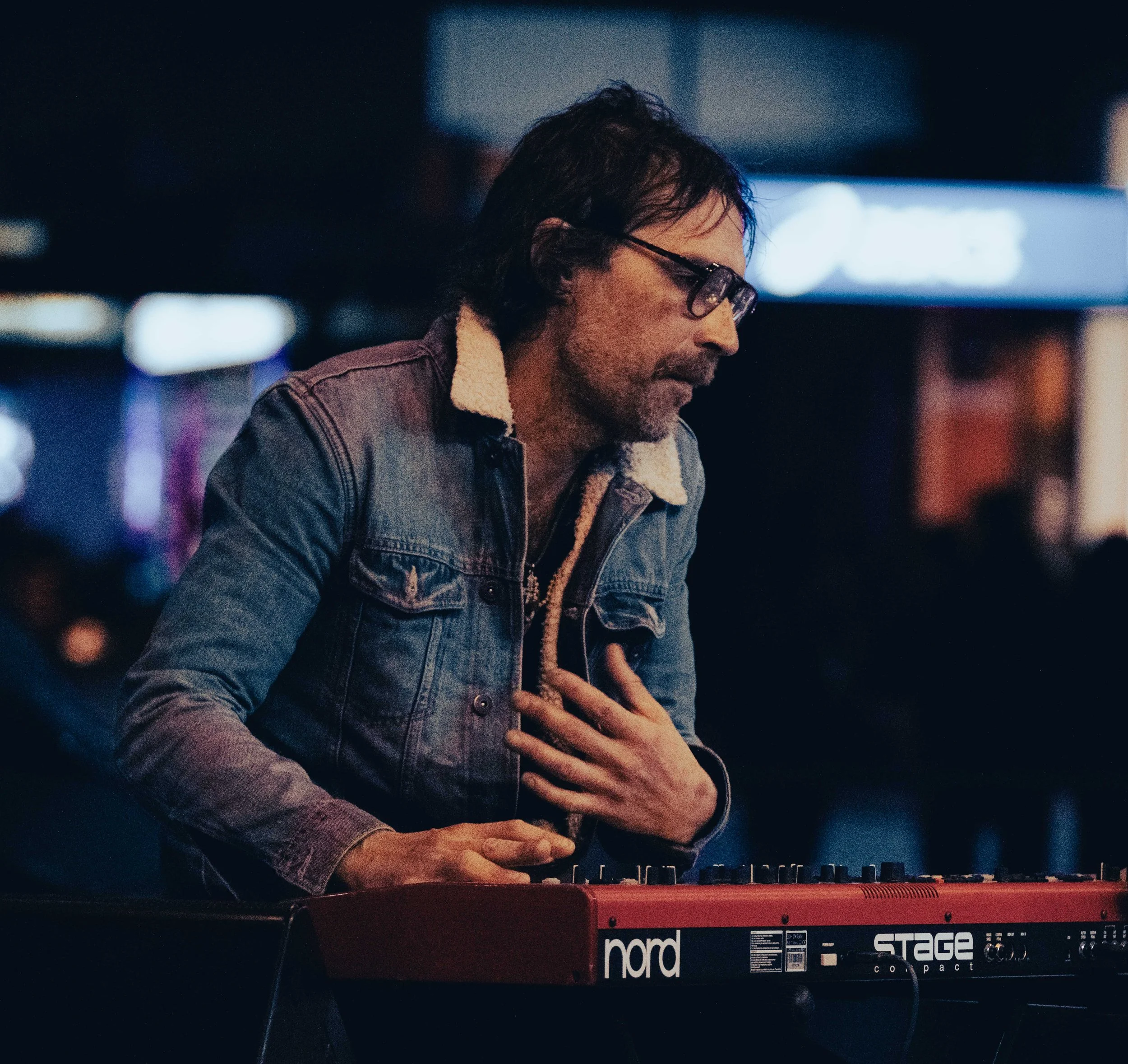 Man with glasses playing a red Nord Stage keyboard in a dark room.