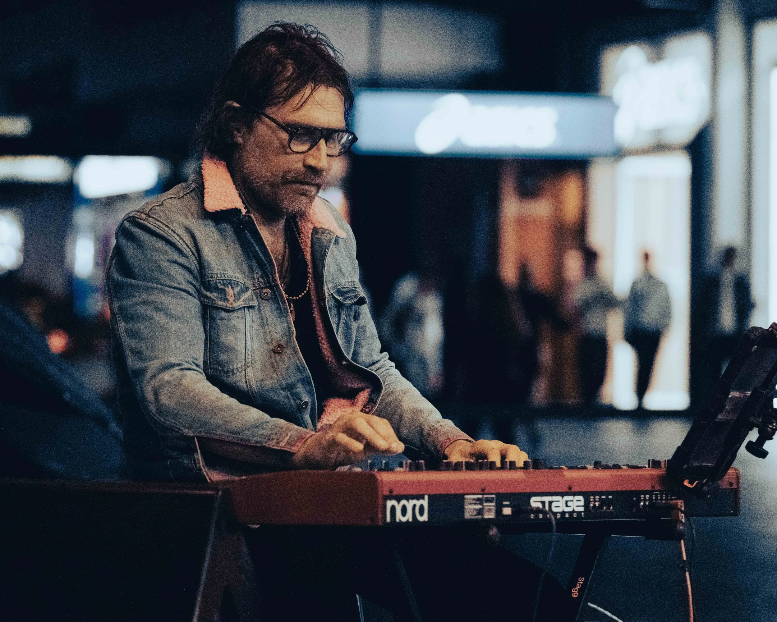 A man with glasses and dark hair playing a red Nord Stage Electronic keyboard in a dimly lit indoor setting.