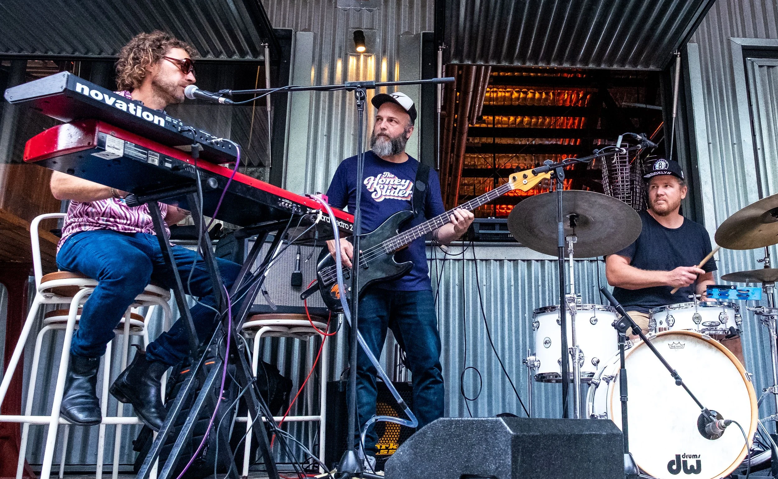 A band performing live with a keyboardist, bassist, and drummer on stage in a rustic setting.