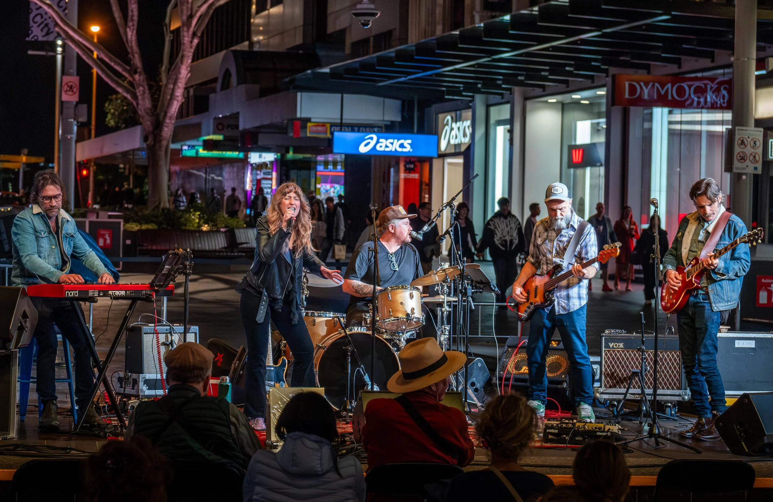 A street musician group performing at night in front of a shopping center. The band includes a woman singing and playing drums, two men playing guitar and bass, and a man on keyboard. Audience members sit on the ground watching the performance.