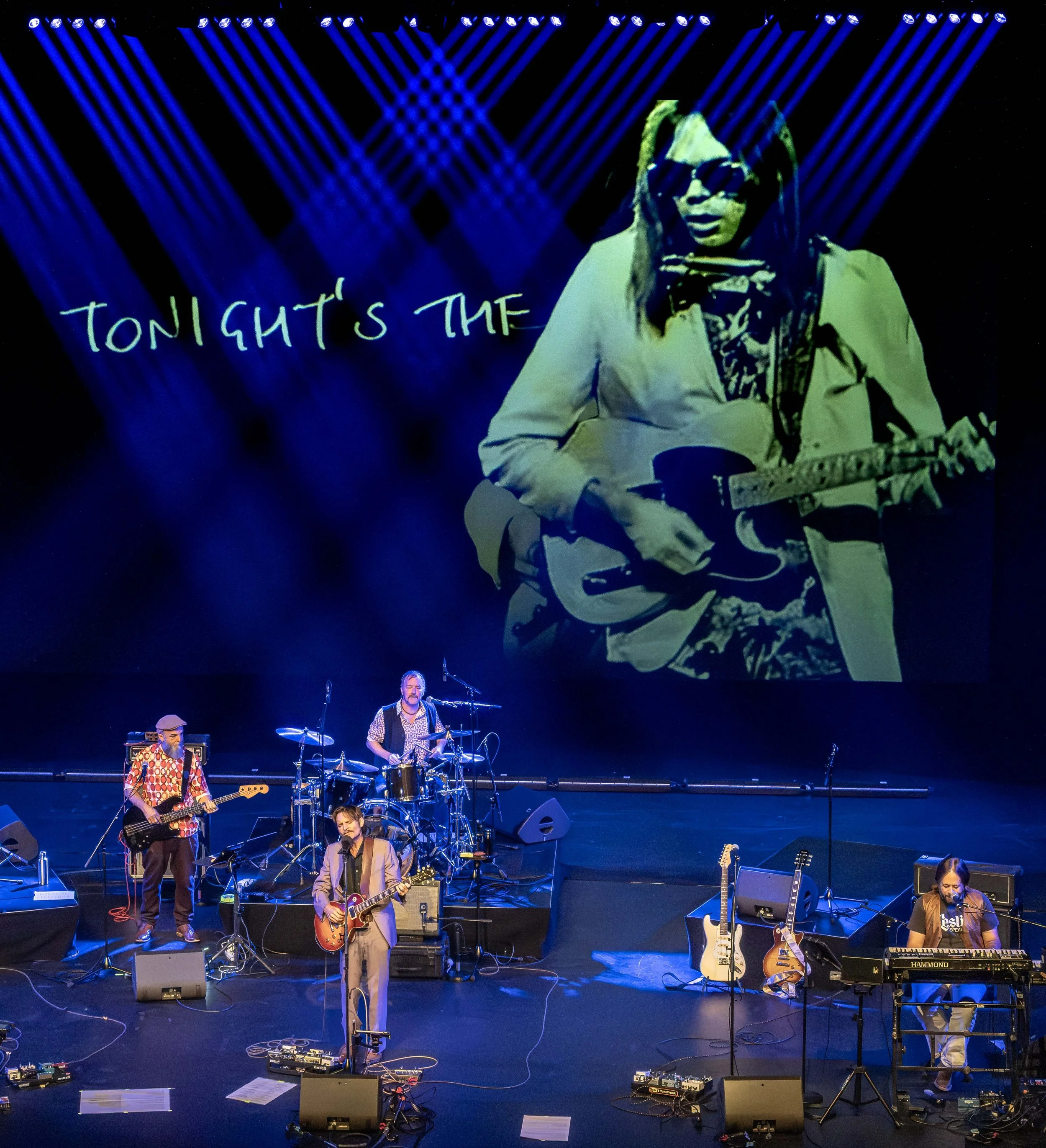 A band performing on stage with a large screen above showing a close-up of a musician in sunglasses playing guitar. The band members are playing various instruments, including guitars, drums, keyboard, and bass, with stage lighting highlighting their