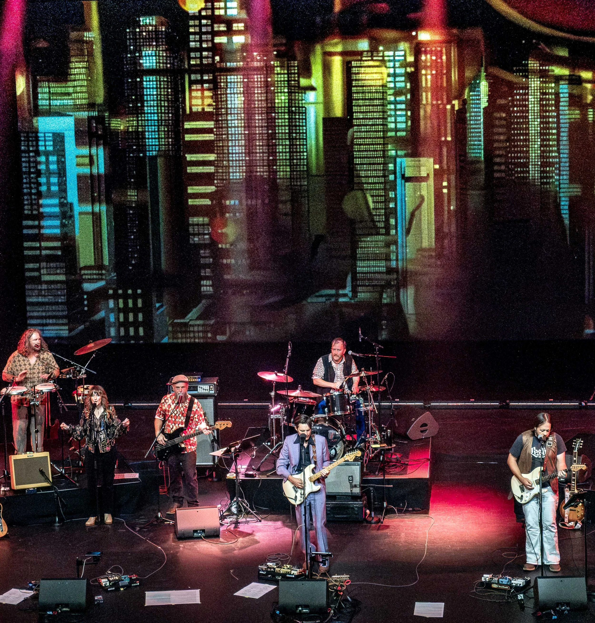 A band performing on stage with a cityscape projection in the background, featuring buildings and city lights. The band includes multiple musicians playing guitars, drums, and singing.