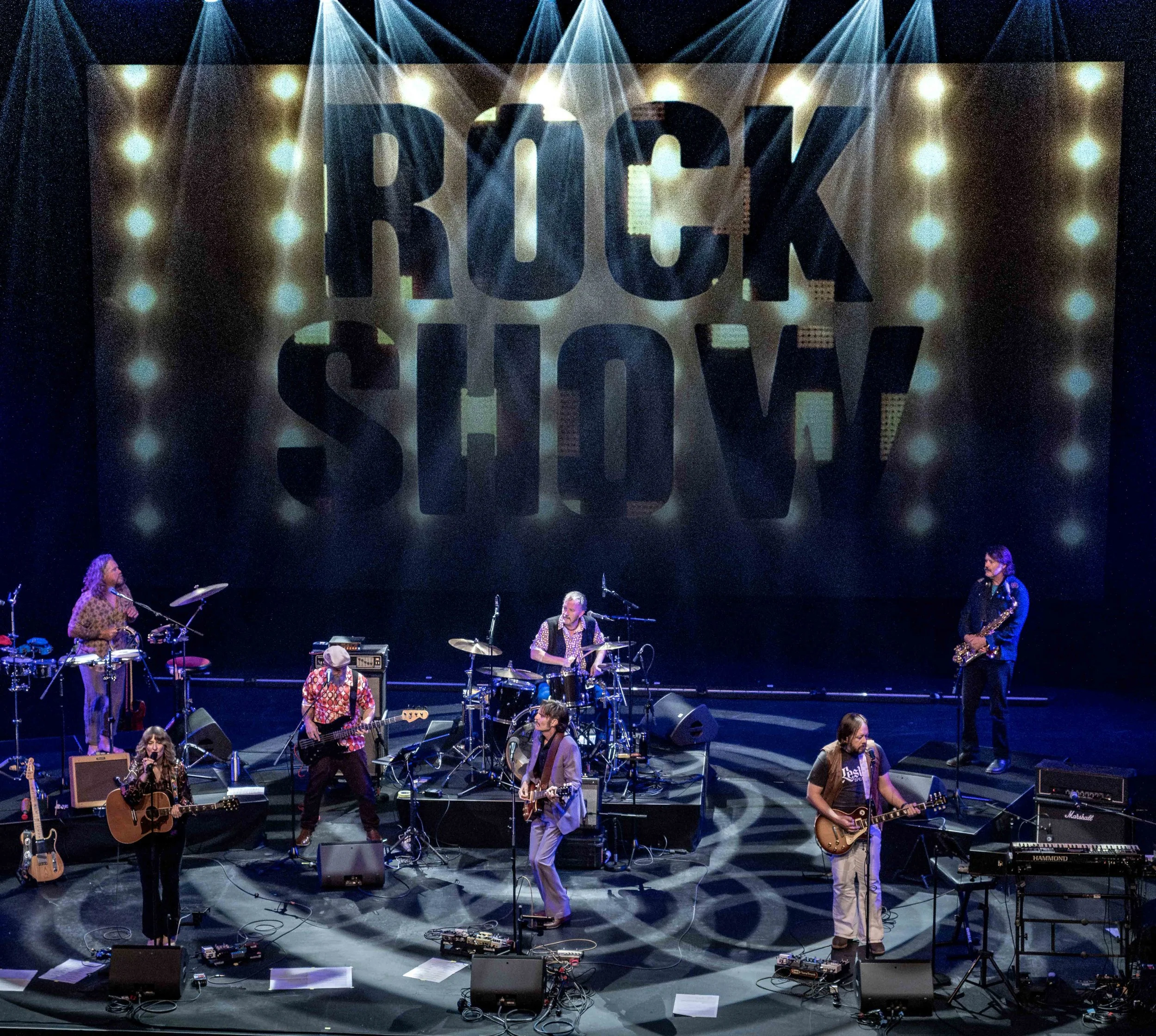 A band performing on stage with multiple musicians playing guitars, drums, and other instruments. A large illuminated sign reading 'ROCK SHOW' is visible behind them.